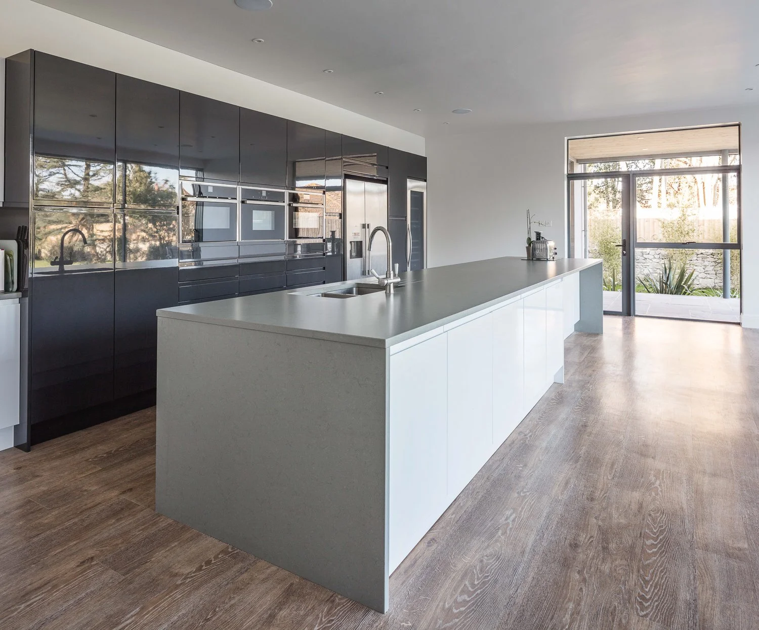 Modern kitchen with white island, black cabinets, wood floor, large glass door, and outdoor view.