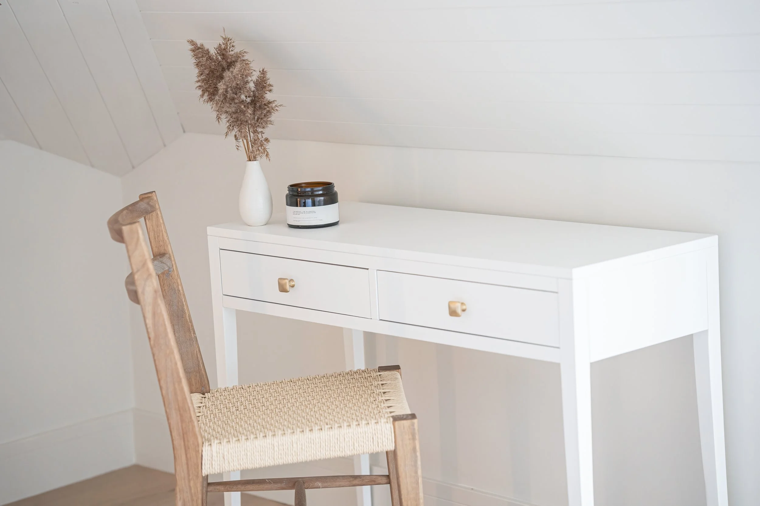 A white desk with two drawers and gold knobs, with a white vase containing dried pampas grass and a black candle jar on top. A wooden chair with woven seat is in front of the desk, in a room with white walls and ceiling.