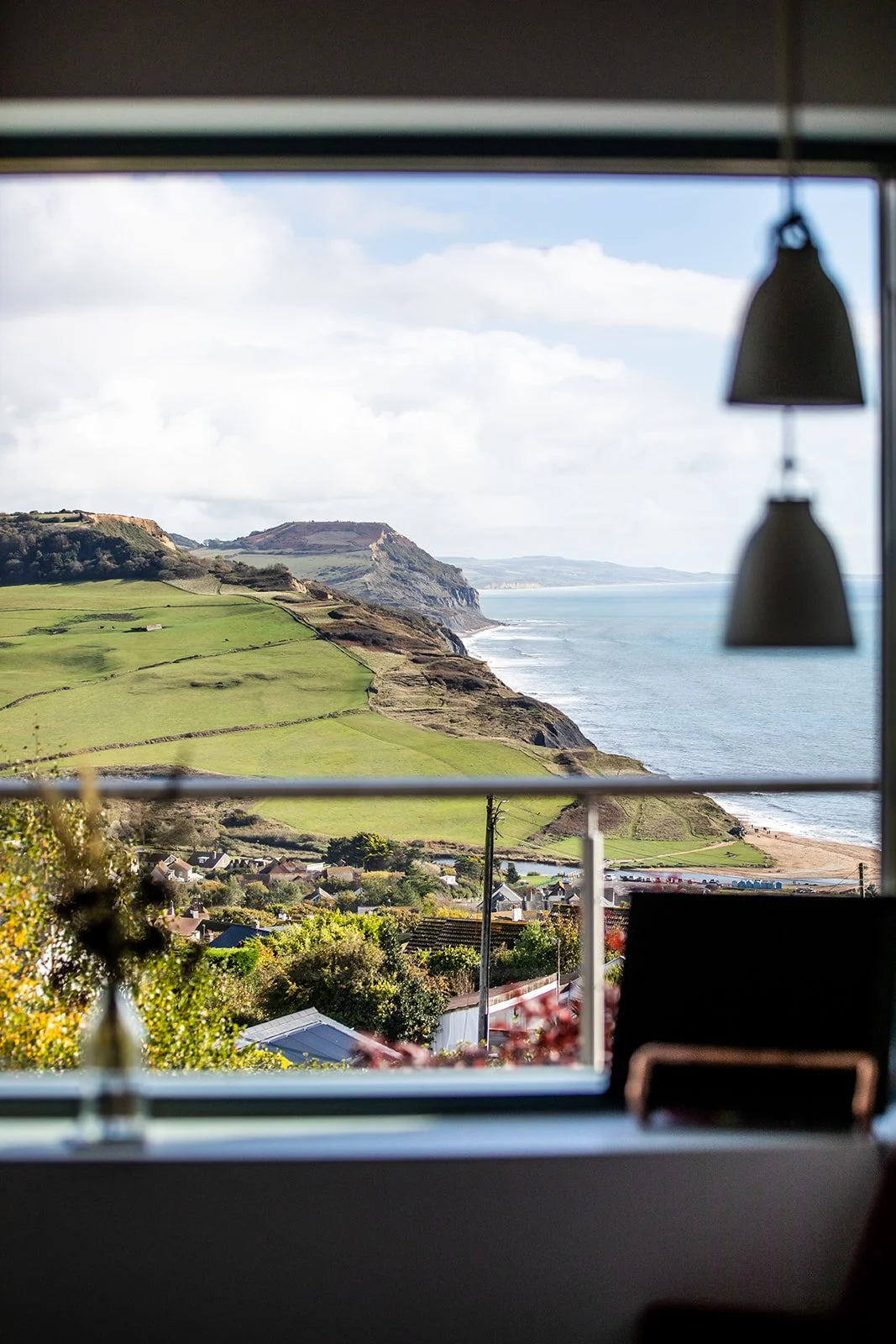 Scenic view of green rolling hills, cliffs, and ocean seen through a window with hanging lamps in the foreground.
