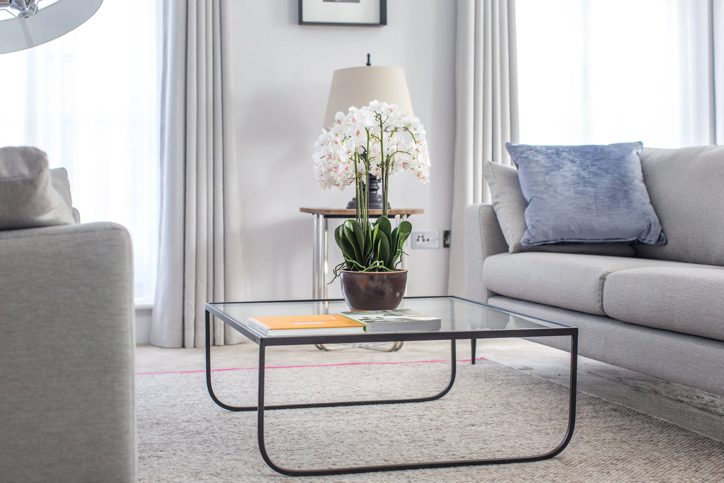 Living room with a glass coffee table holding a potted white orchid, beige sofas with blue pillows, a side table with a lamp, and large windows with white curtains.