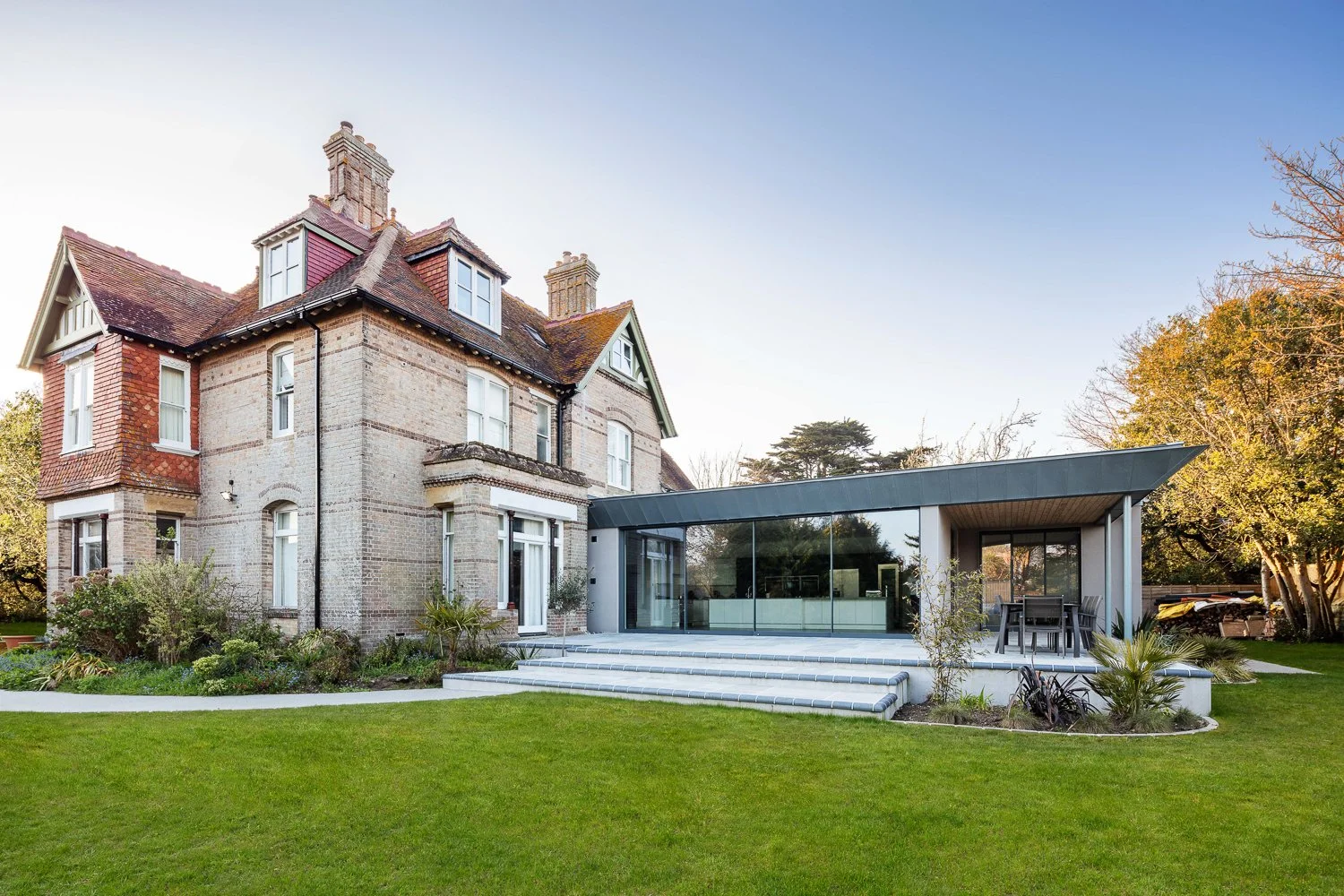 A large house with a brick and stone exterior, featuring a modern glass extension with an outdoor dining area on a multi-step patio, surrounded by a lush green lawn and trees.