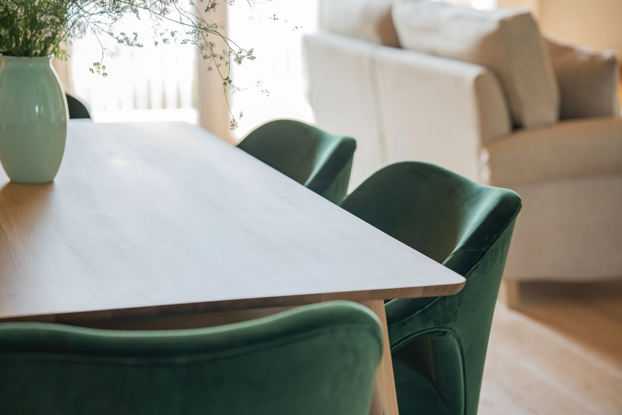 A dining room table with green velvet chairs and a white vase with green plants on a wooden table, sunlight coming through windows.