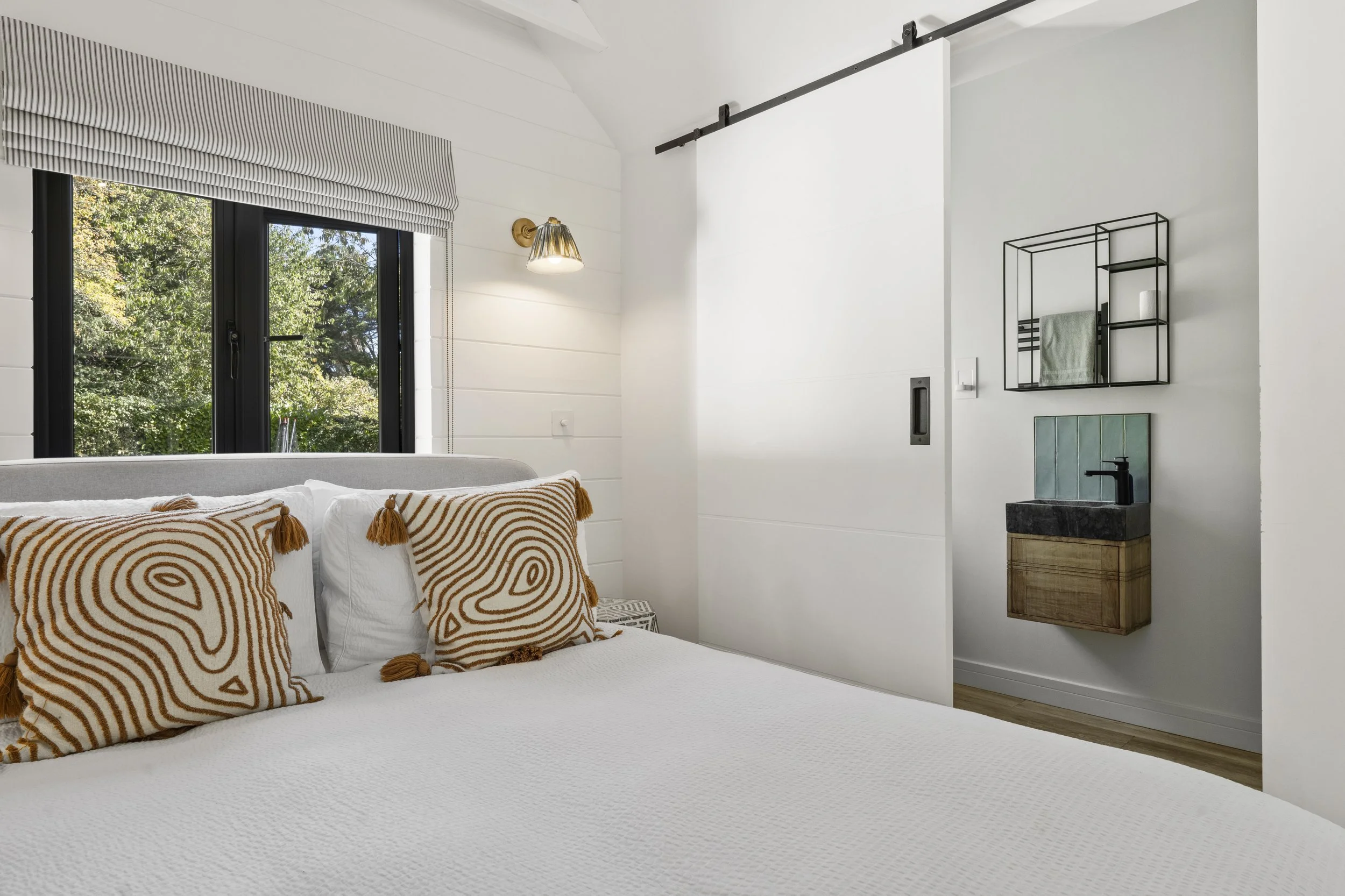 Bedroom with white walls, a bed with white bedding and decorative pillows, a window with trees outside, a wall-mounted light, and a sliding door leading to a small washbasin area.