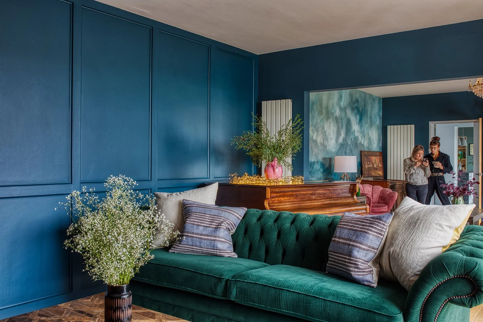 Living room with a dark green velvet sofa, pillows, and wall panels, a wooden console table with a pink vase of greenery, and two women standing in the background near a doorway.