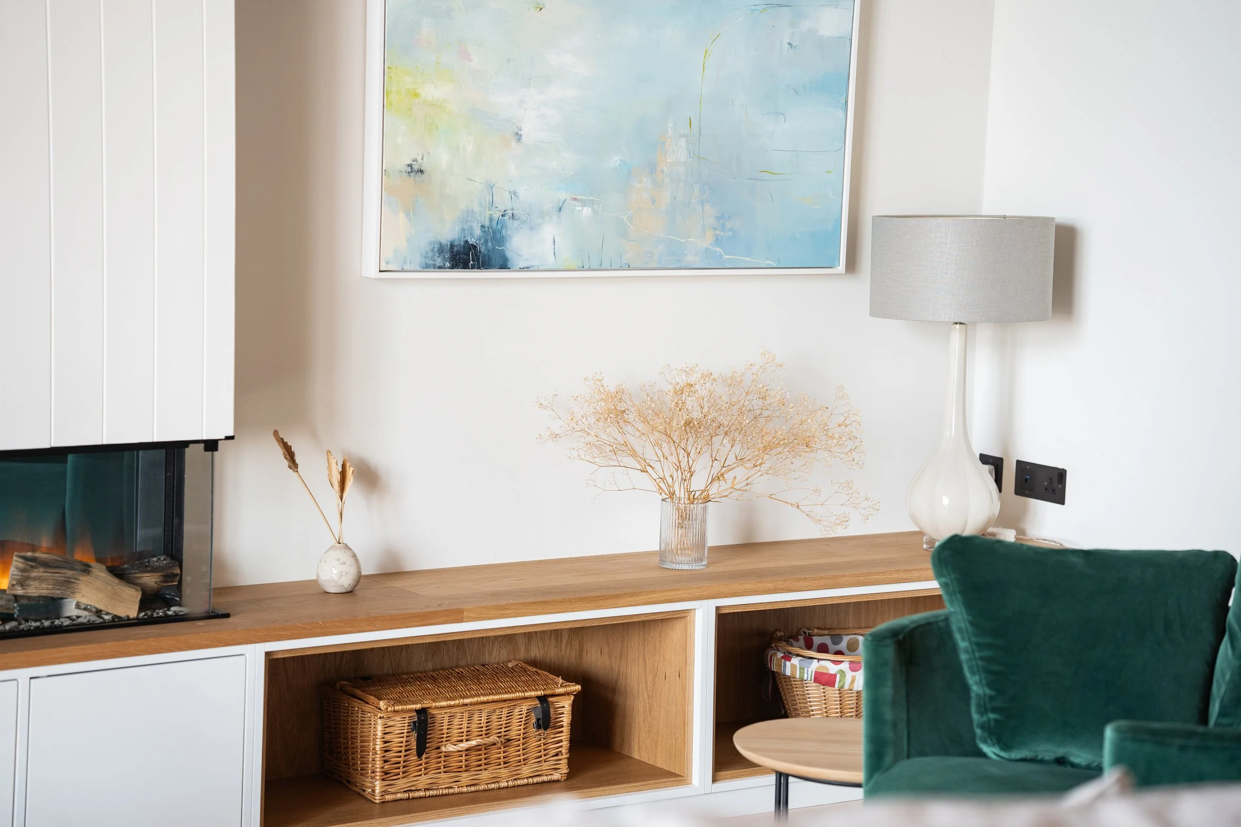 Living room with a modern wood and white TV console, abstract art on the wall, a table lamp with a gray shade, a glass vase with dried flowers, a small wicker basket, and a green velvet armchair.