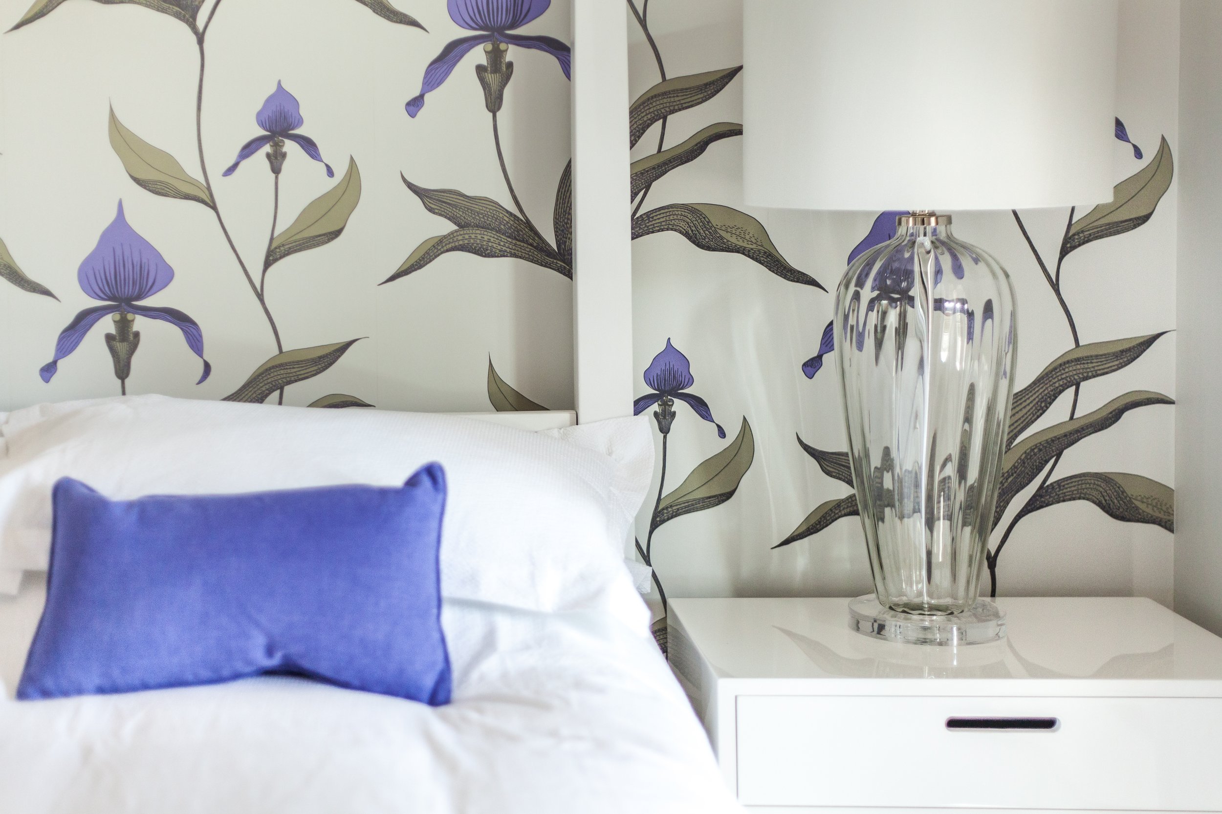 Close-up of a white bed with white pillows, one of which is accented with a blue pillow, next to a white nightstand with a large glass table lamp and floral wallpaper featuring purple flowers and green and brown leaves.