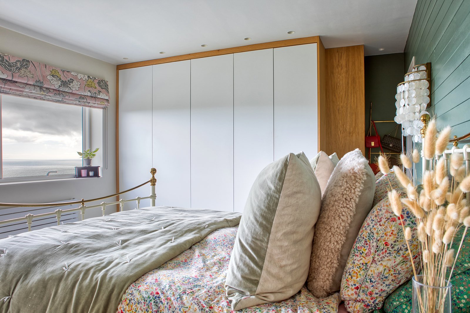 A cozy bedroom with a bed, decorative pillows, a window with a floral valance showing a cloudy sky and ocean view, white built-in closet, a teal wall with a modern white and gold lamp, and a vase of dried grasses.