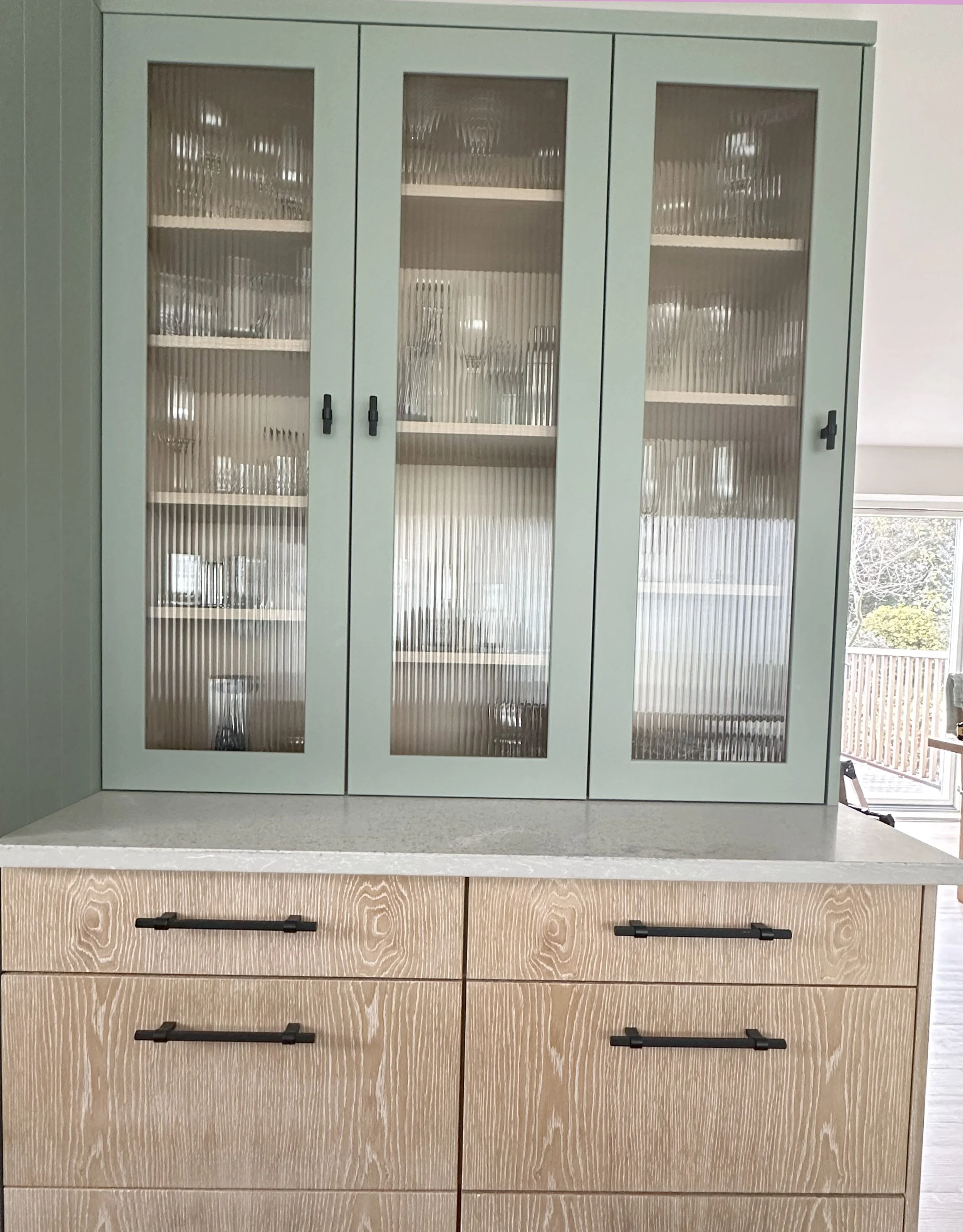 A kitchen cabinet with glass doors containing glassware, placed above a wooden dresser with six drawers and black handles.
