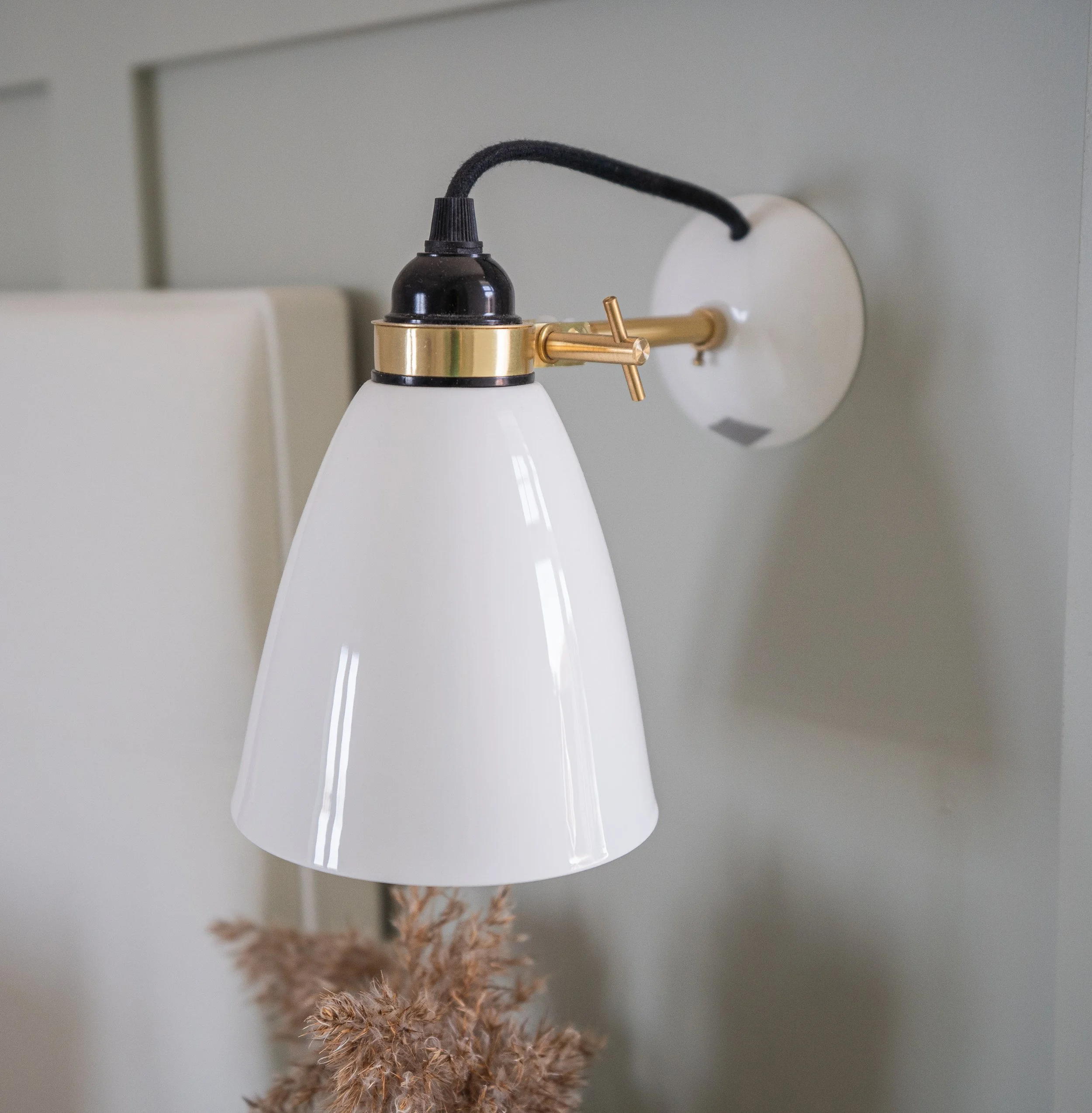 Wall-mounted lamp with a white conical shade, black and gold accents, and a textured black cord, positioned above dried pampas grass.