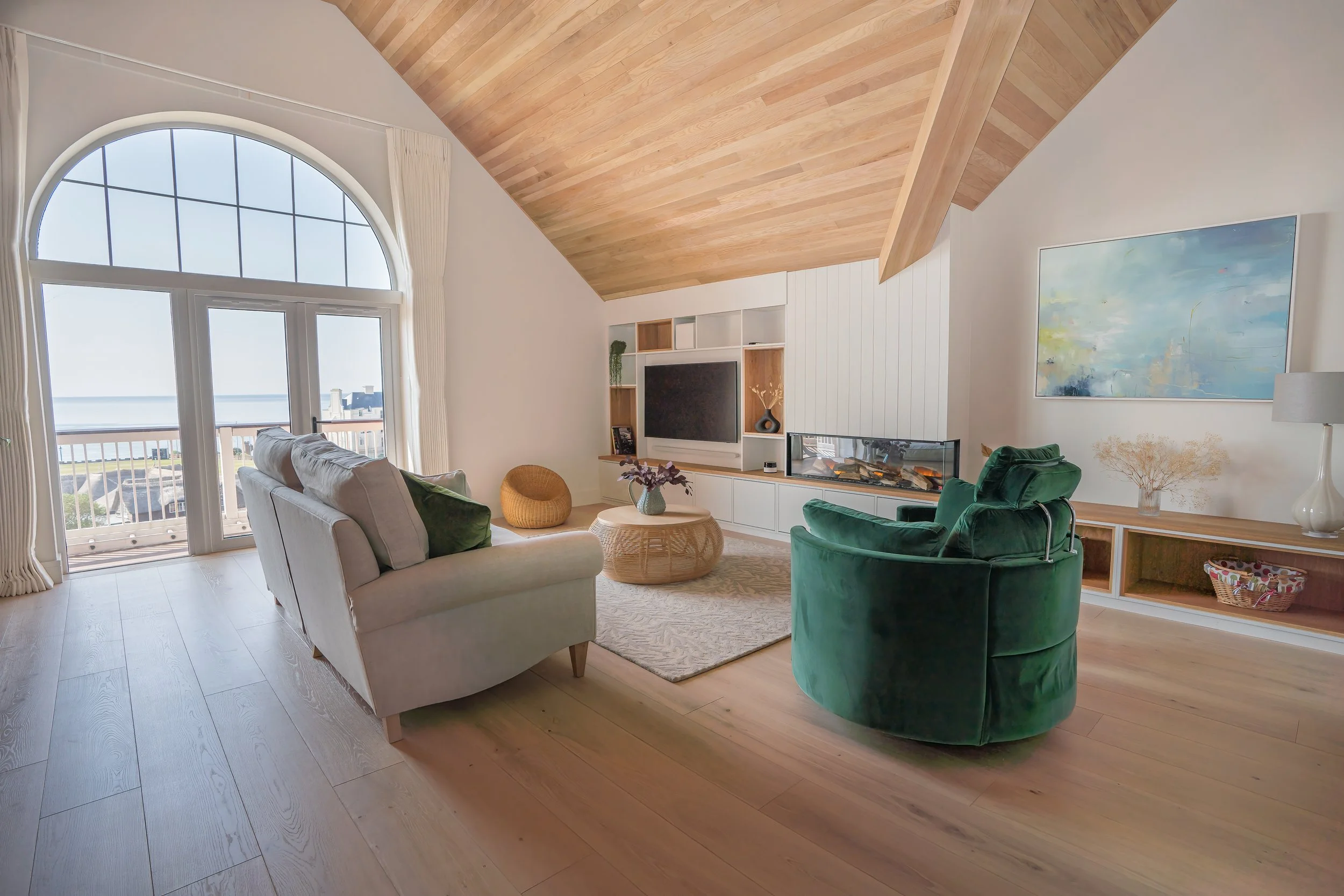 Living room with large arched window, white sofa, green velvet armchair, coffee table with vase, built-in wall unit with a TV and fireplace, wooden flooring and ceiling, abstract artwork on wall, with view of the outdoors.
