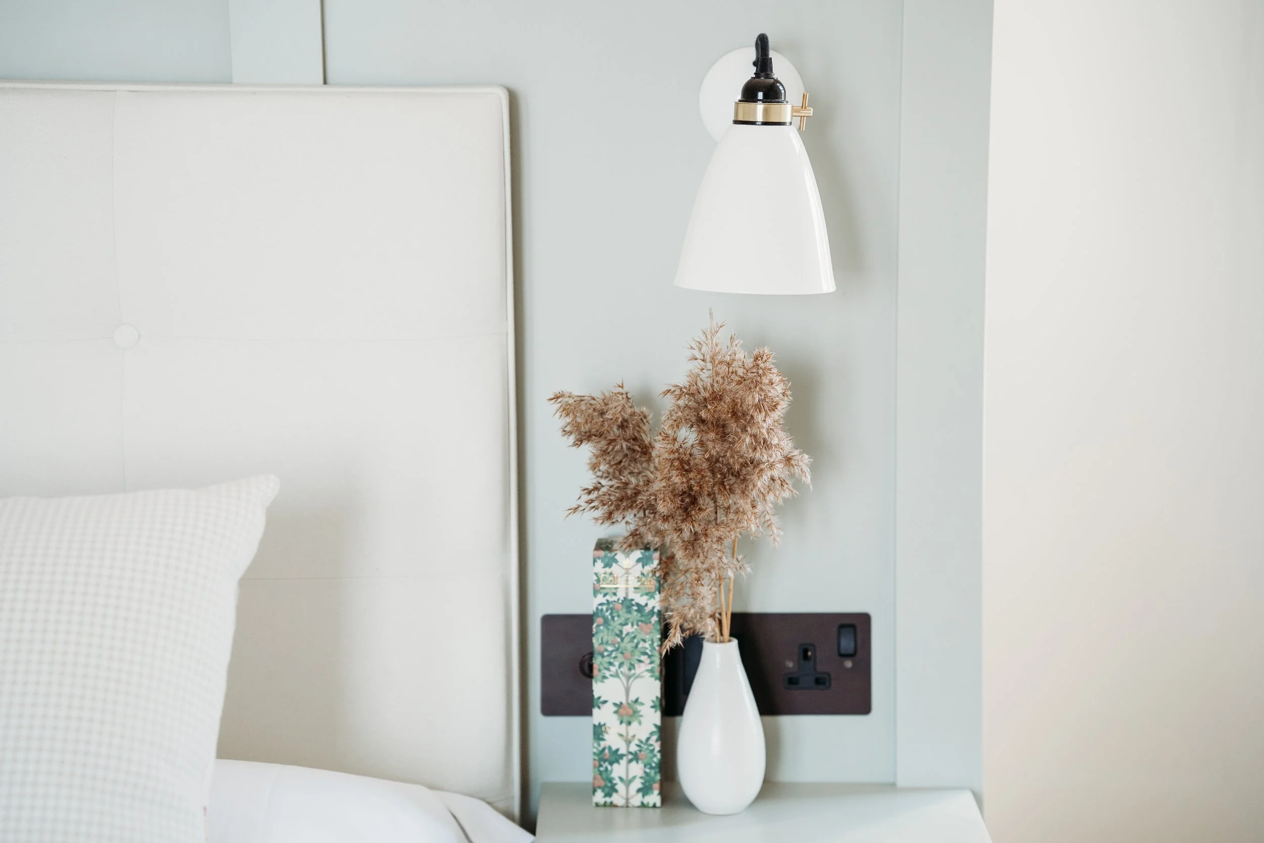 A bedroom nightstand with a white vase holding dried pinkish pampas grass, a decorative box with floral pattern, a white wall-mounted lamp, electrical outlets, and part of a bed with white bedding and a textured pillow.