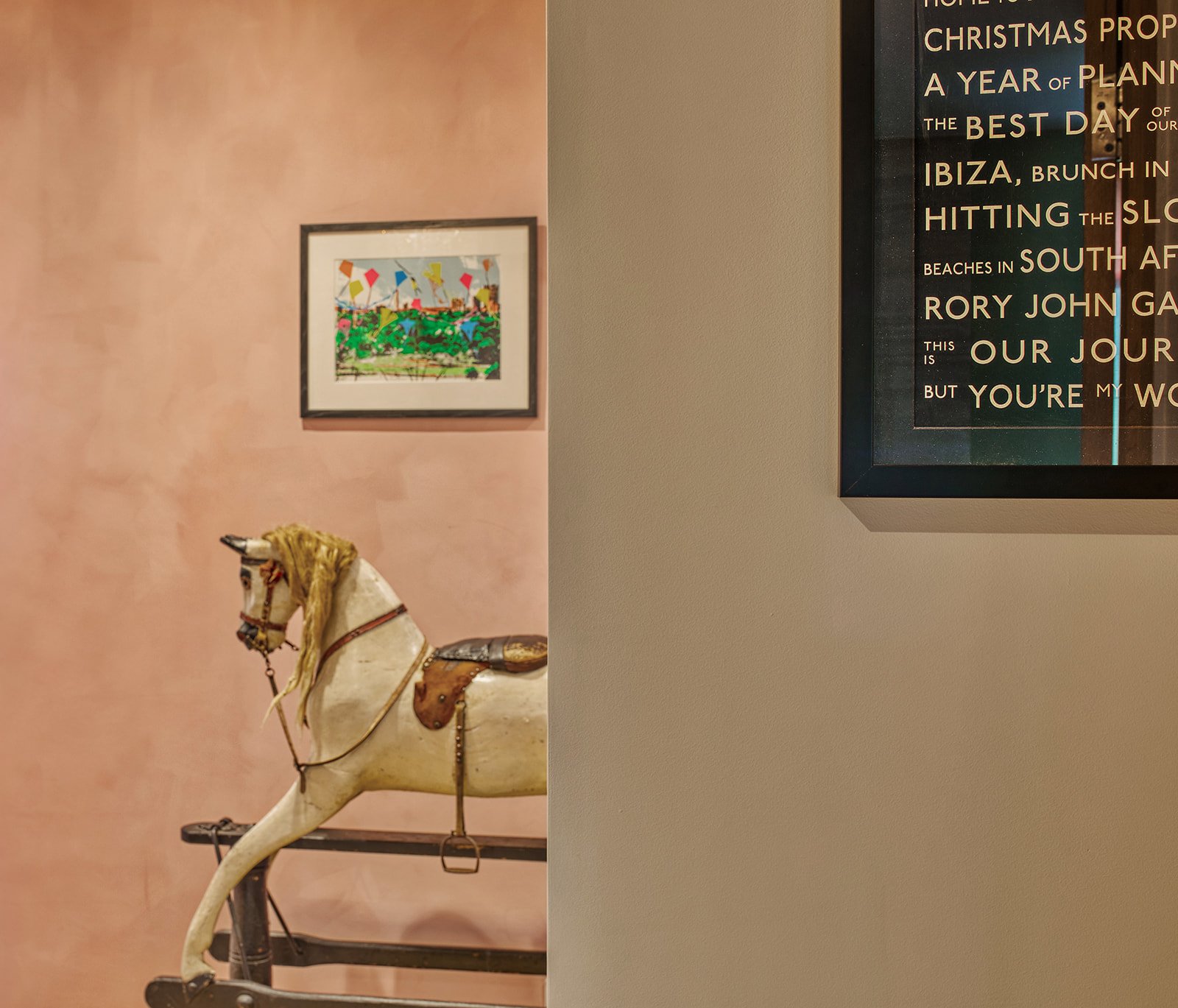 A vintage rocking horse with a white body, a golden mane, and a saddle, standing in front of a pink textured wall with a colorful framed artwork of a landscape hanging above it. To the right, part of a framed poster with text about travel and holiday