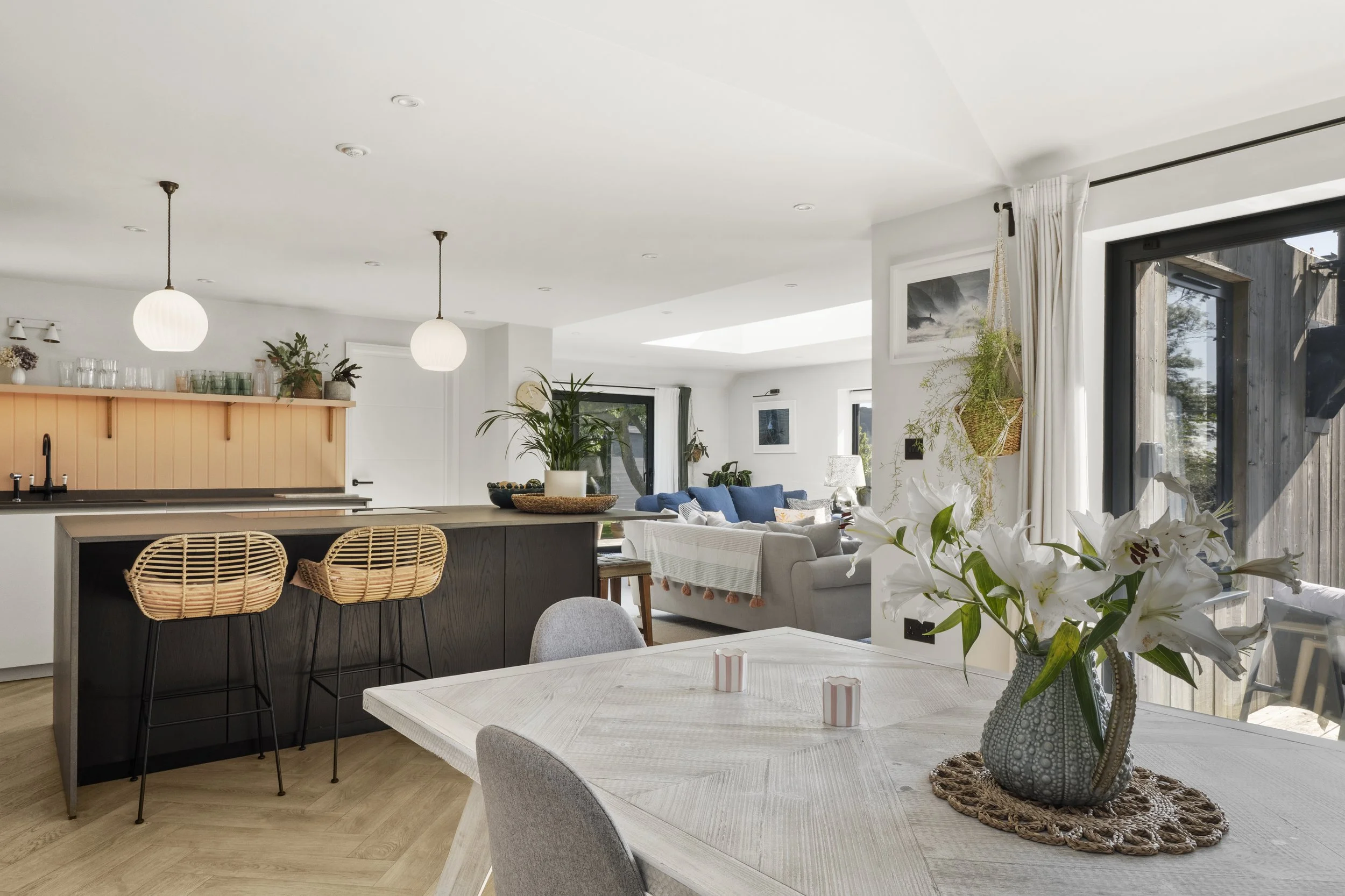 Bright open-concept living space featuring a dining area with a white table and a vase of white lilies, a kitchen with a black island and two wicker barstools, and a cozy seating area with a white sofa decorated with blue cushions and a throw, large 
