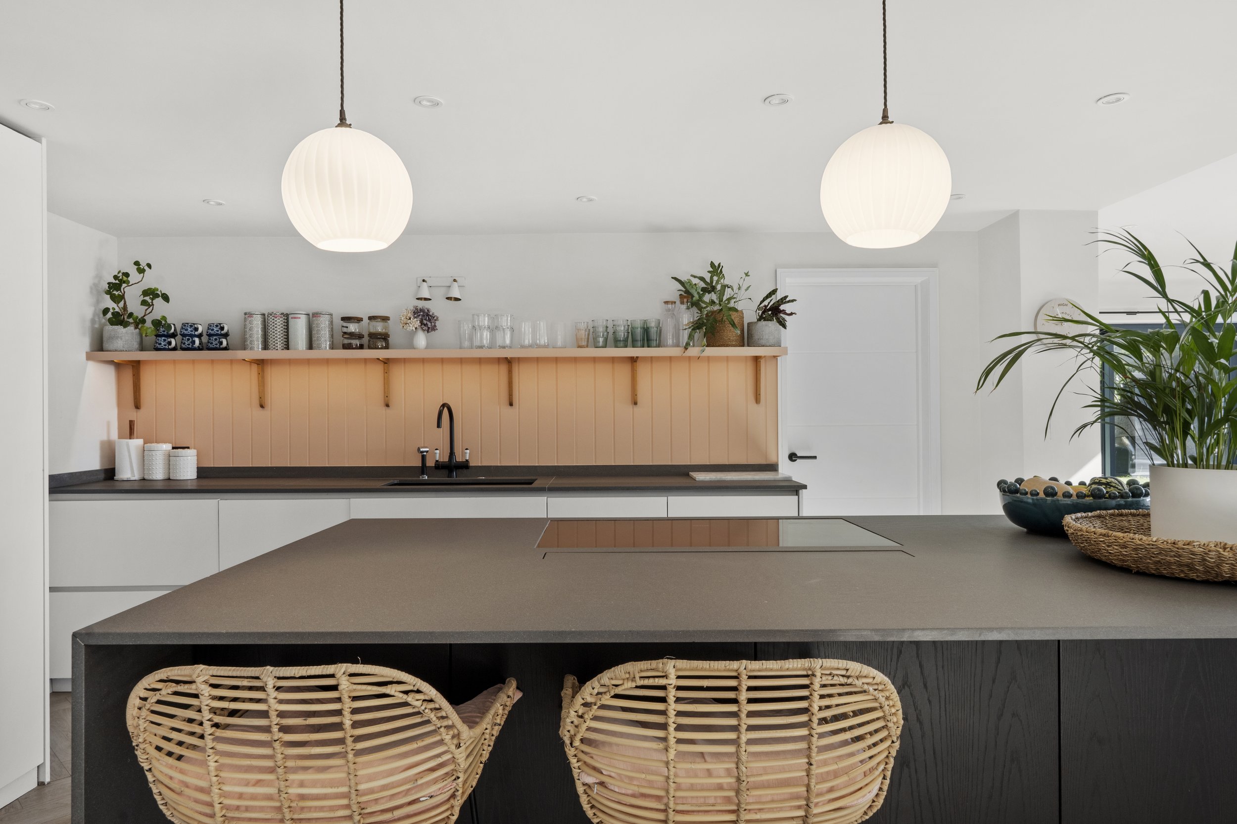 Modern kitchen with white cabinets, black countertop, and peach backsplash. Two pendant lights hang from the ceiling, and there are green plants and decorative items on the shelves. Two wicker bar stools are at the island, and a bowl with decorative 