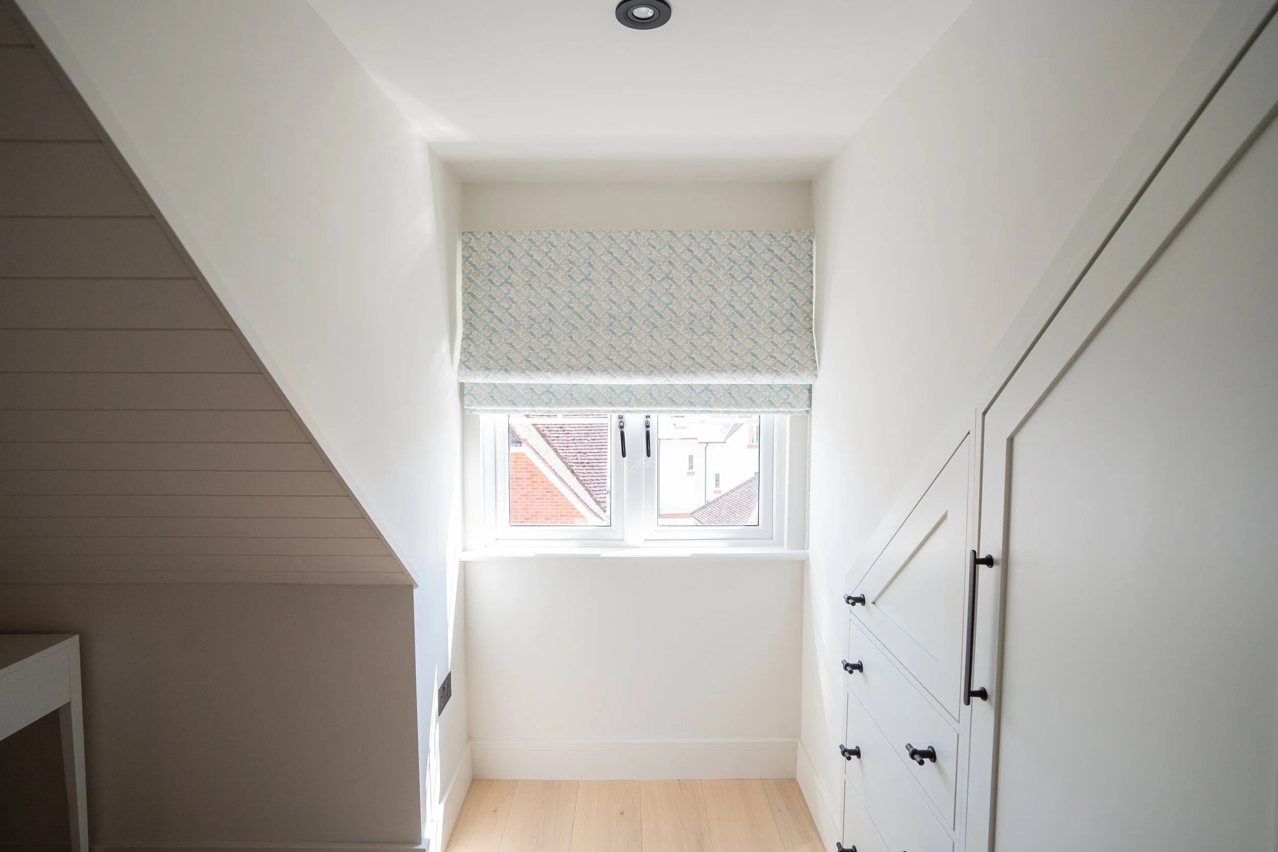 Bright corner of a room with a window, blue patterned window shade, white walls, sloped ceiling, and built-in white cabinetry.
