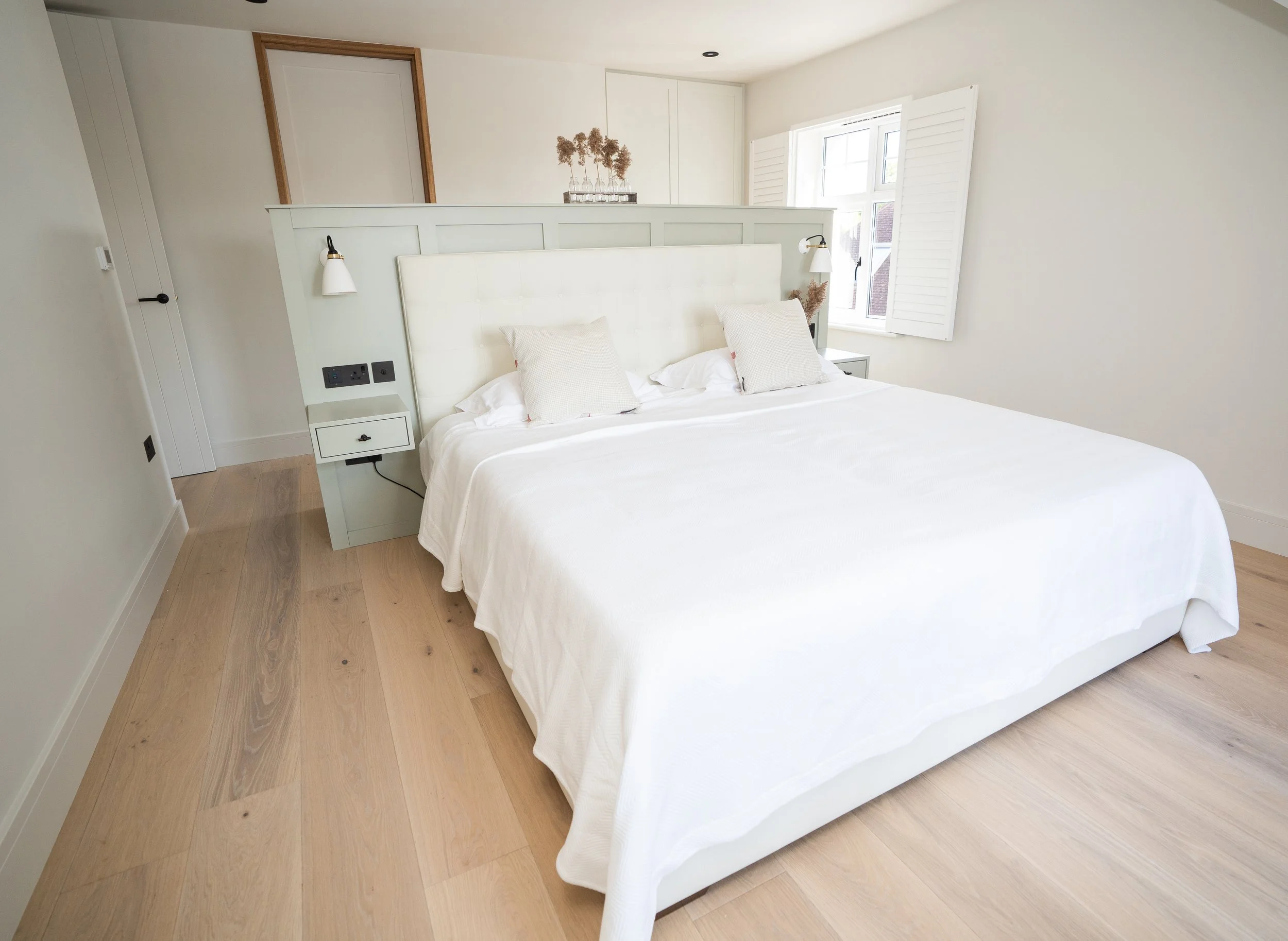 A bright bedroom with a large bed, white bedding, and two beige pillows. There are wall-mounted lamps on each side of the bed, a small nightstand on the right, and a window with white shutters on the right side. The room has light wood flooring and m