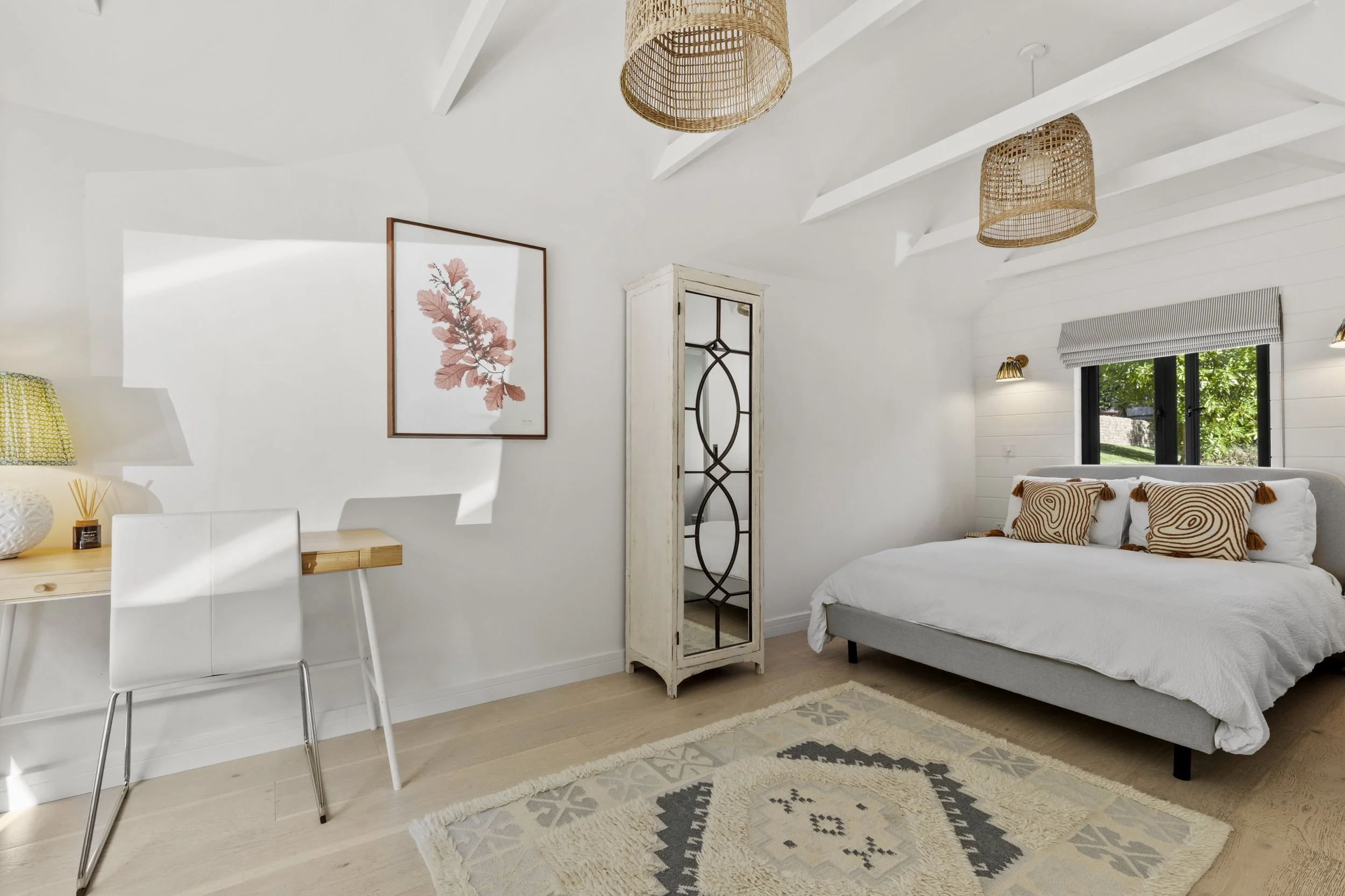 Bright bedroom with white walls, vaulted ceiling with exposed beams, a gray bed with pillows, a window with greenery outside, a white wooden wardrobe with glass door, a small desk with a white chair, a framed pink floral art print, woven pendant ligh