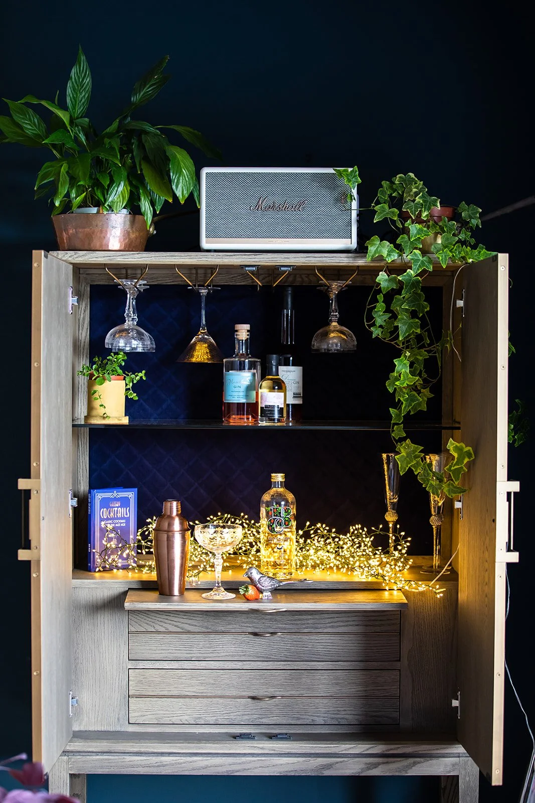 A wooden bar cabinet with plants on top, wine glasses hanging upside down, various bottles of alcohol and drinkware inside, decorative string lights, and cocktail-related items.