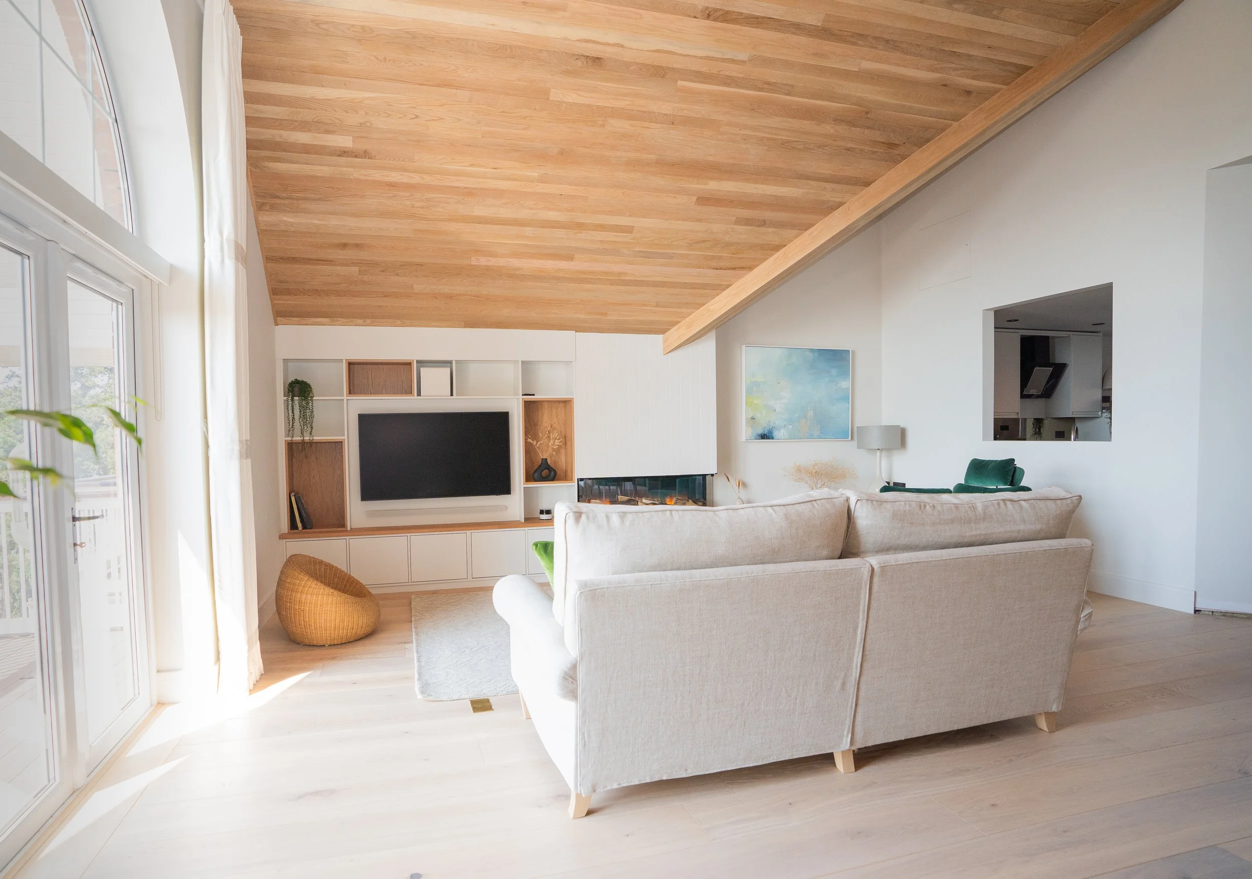Living room with beige sofa, white bookshelf with TV, wooden ceiling, large window, abstract painting, lamp, and other furniture.