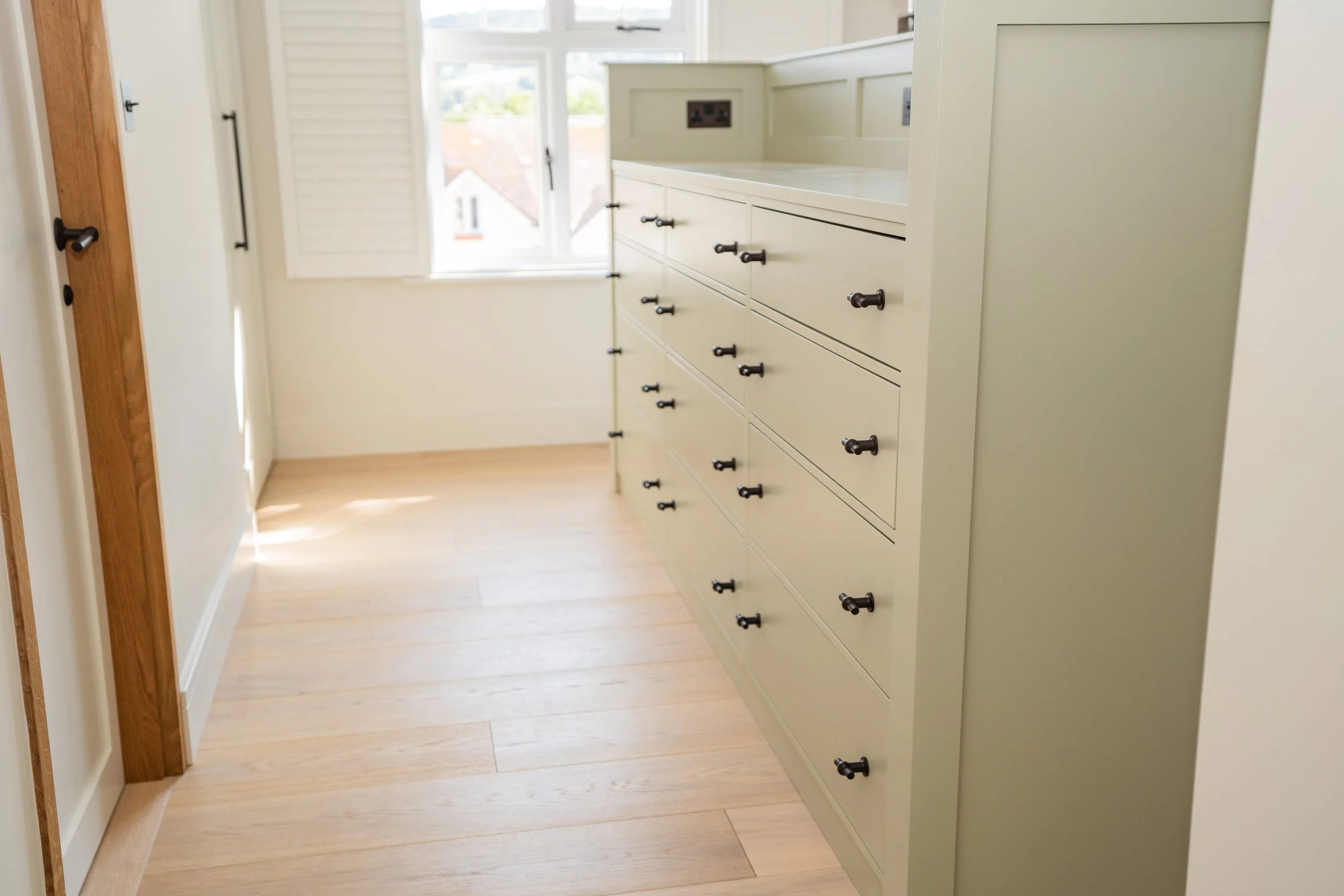 A cream-colored dresser with black knobs in a sunlit room with hardwood floors and a window with white shutters.