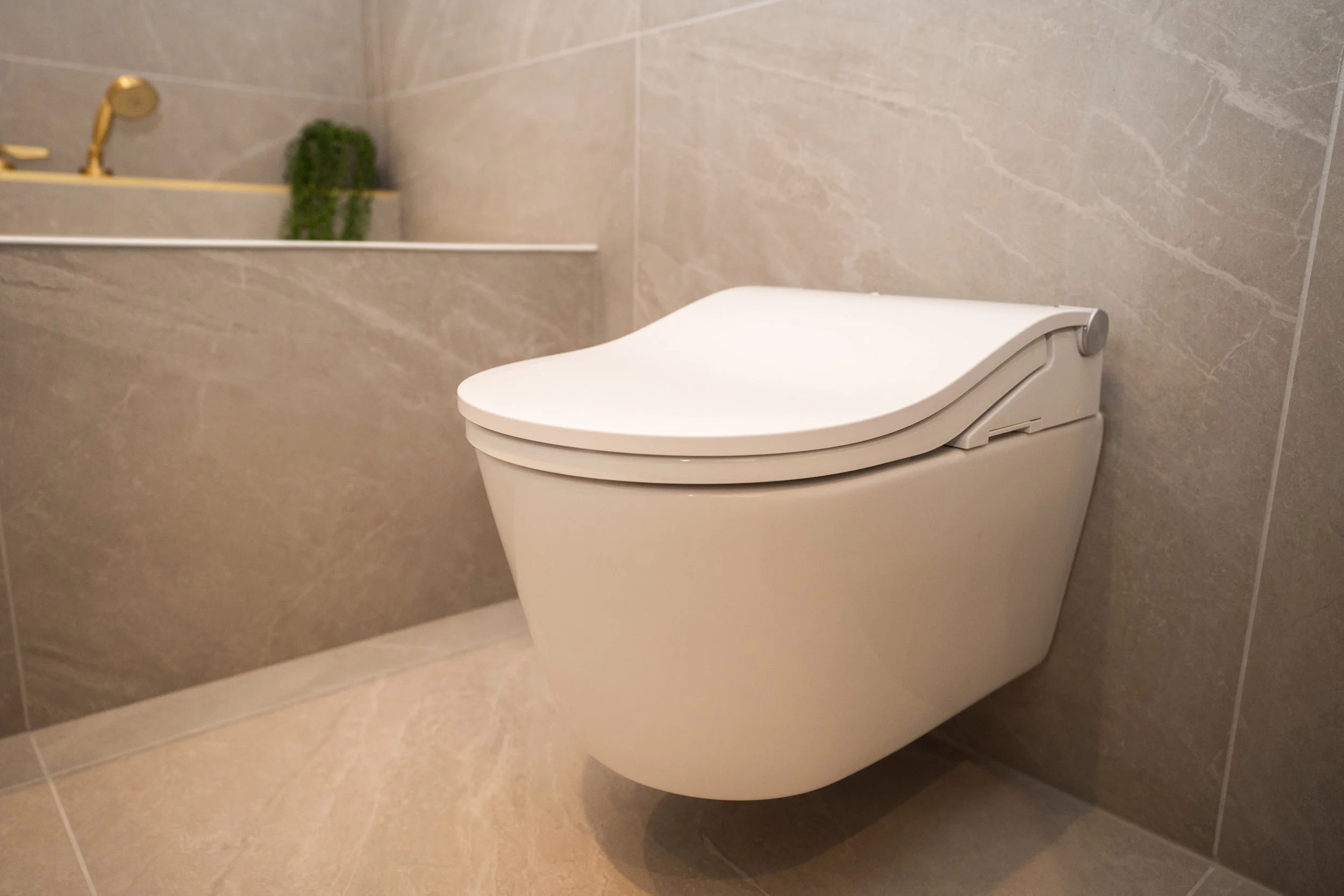A modern, wall-mounted, white toilet in a bathroom with beige tiled walls and floor, with a gold faucet and a small potted plant in the background.