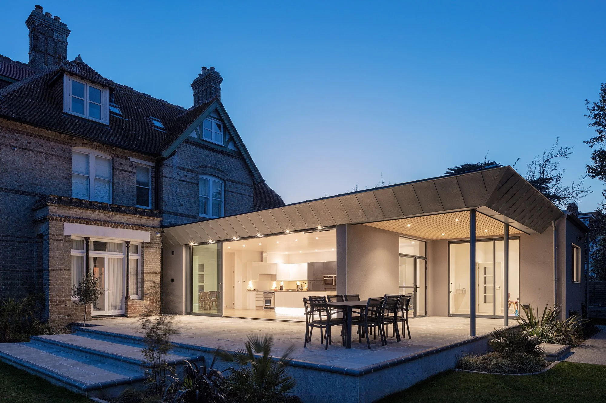 A modern house extension with large glass sliding doors, an outdoor dining table with chairs, and a built-in pool with landscaping in the evening.