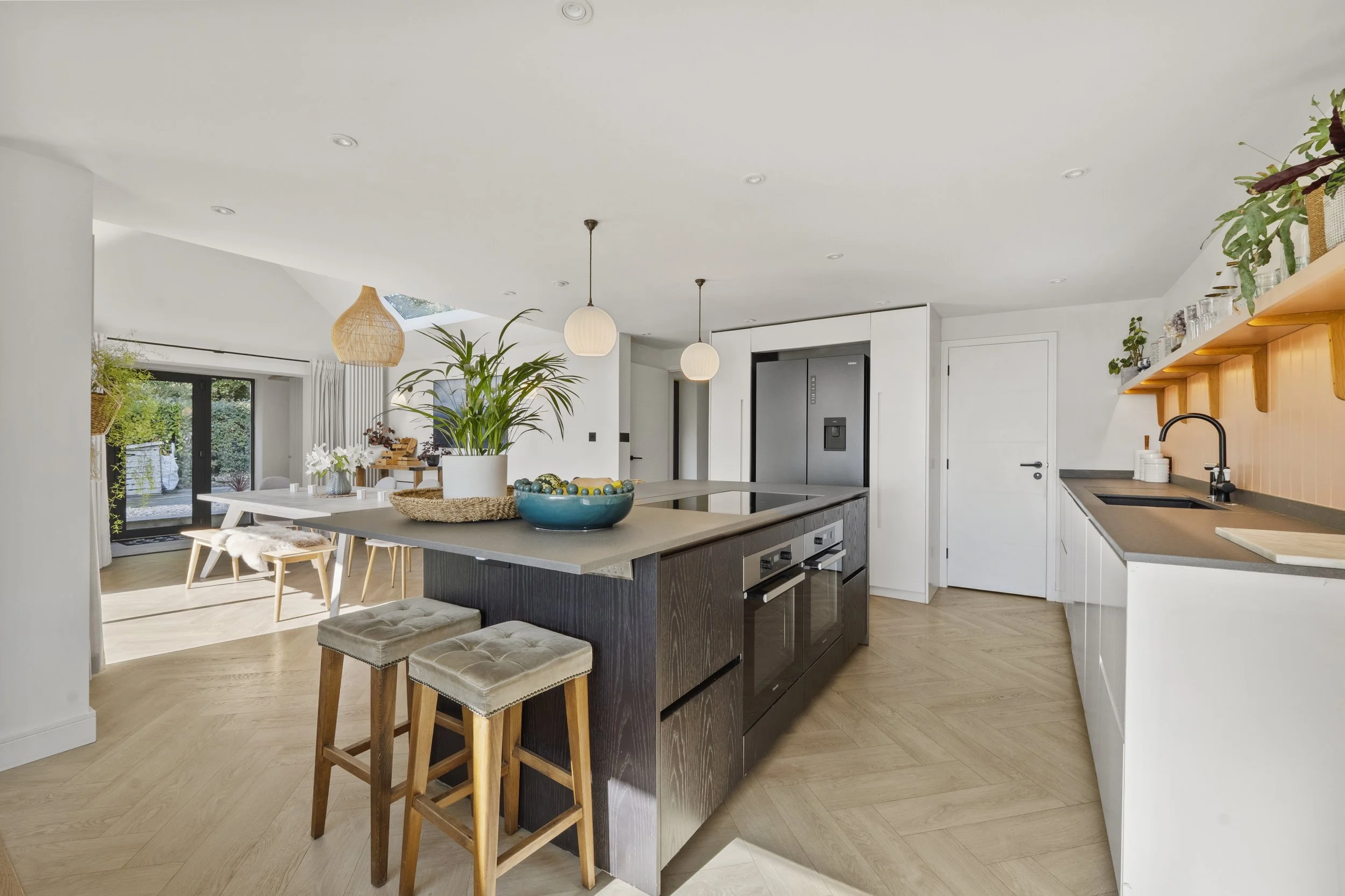 Modern kitchen with an island, dark lower cabinets, white upper cabinets, hanging pendant lights, and dining area visible through sliding glass doors.