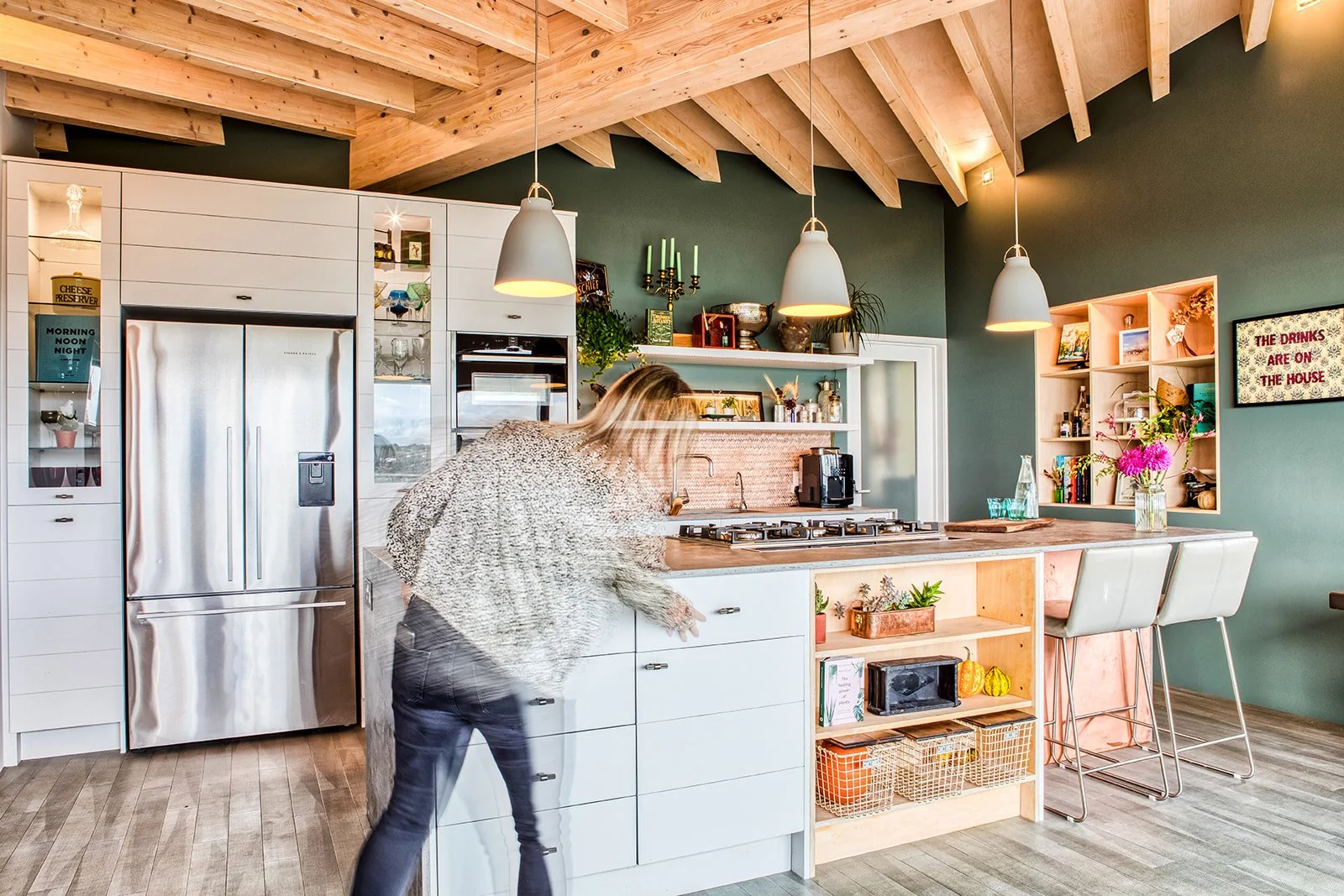 A modern kitchen with wooden vaulted ceiling, white cabinetry, a refrigerator, open shelves with various decor items, and a kitchen island with barstools. A woman is leaning over the island. Pendant lights hang from the ceiling.