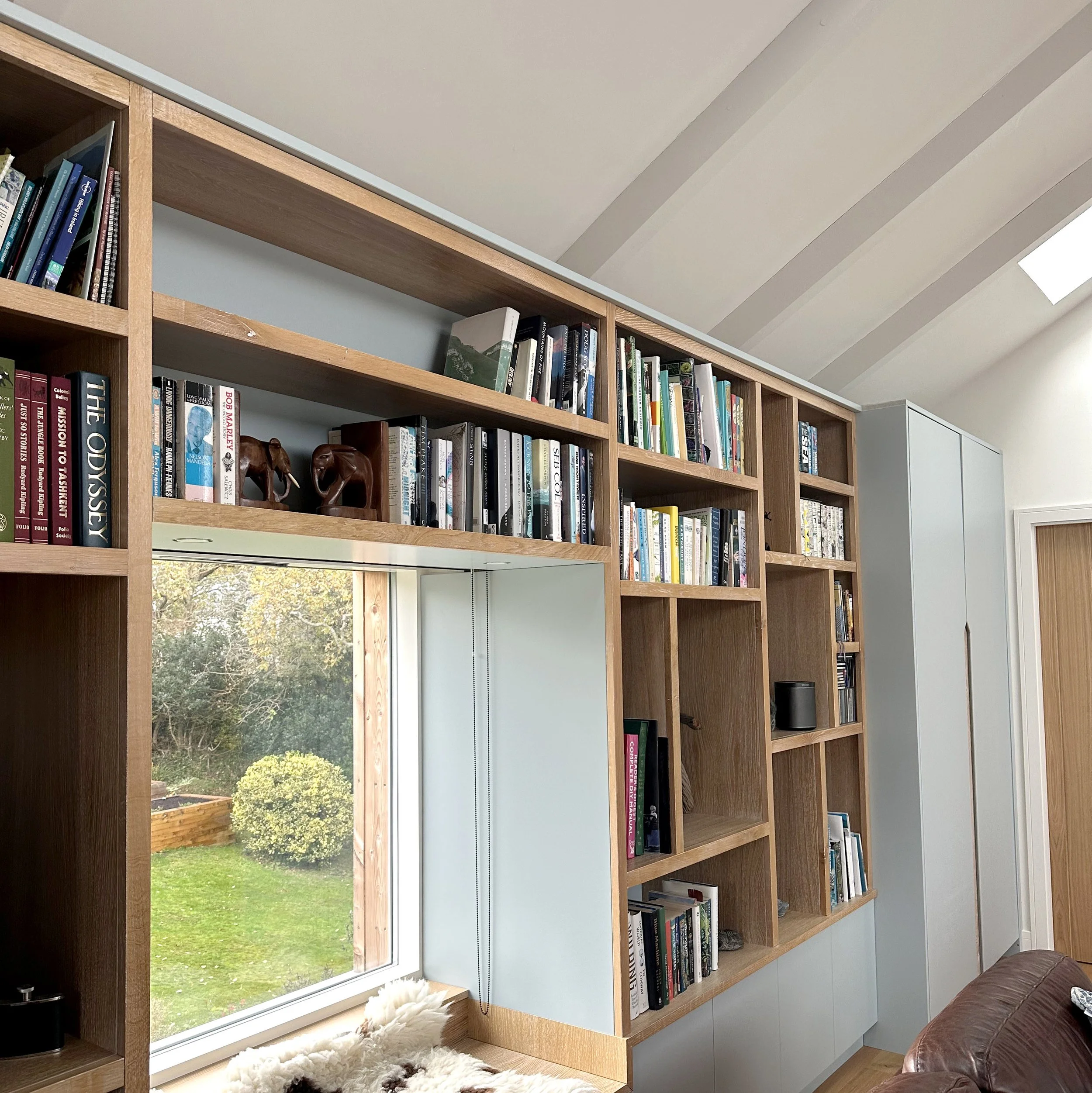 A wooden bookshelf with multiple sections filled with books, located next to a window with a view of a green garden. The room has a relaxed, cozy atmosphere with a fluffy rug near the window and a brown leather sofa partially visible on the right.