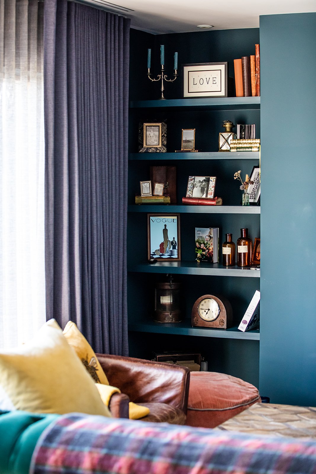 A view of a living room corner with a blue bookshelf filled with framed photos, books, and decorative items. Next to the bookshelf is a window with blue curtains. In the foreground, part of a sofa with yellow and patterned pillows is visible.