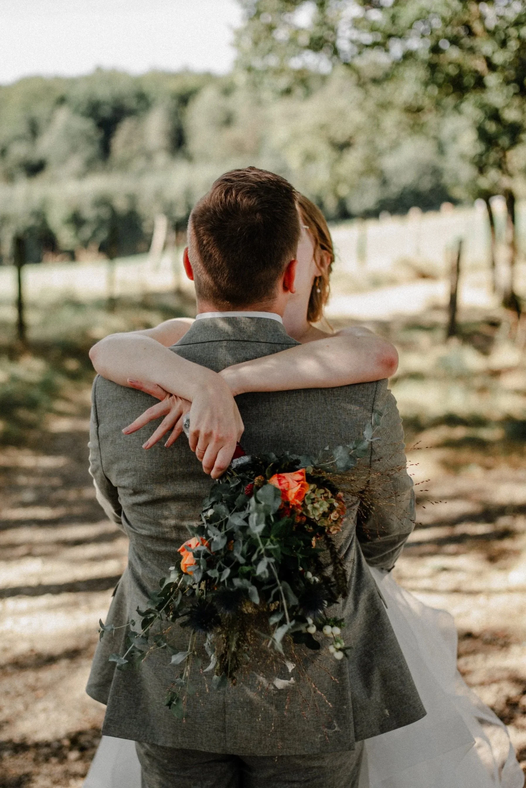 Ein verliebtes Paar, das sich umarmt, im Freien bei Sonnenlicht, mit einem Blumenstrauß in der Hand, wahrscheinlich bei einer Hochzeit.