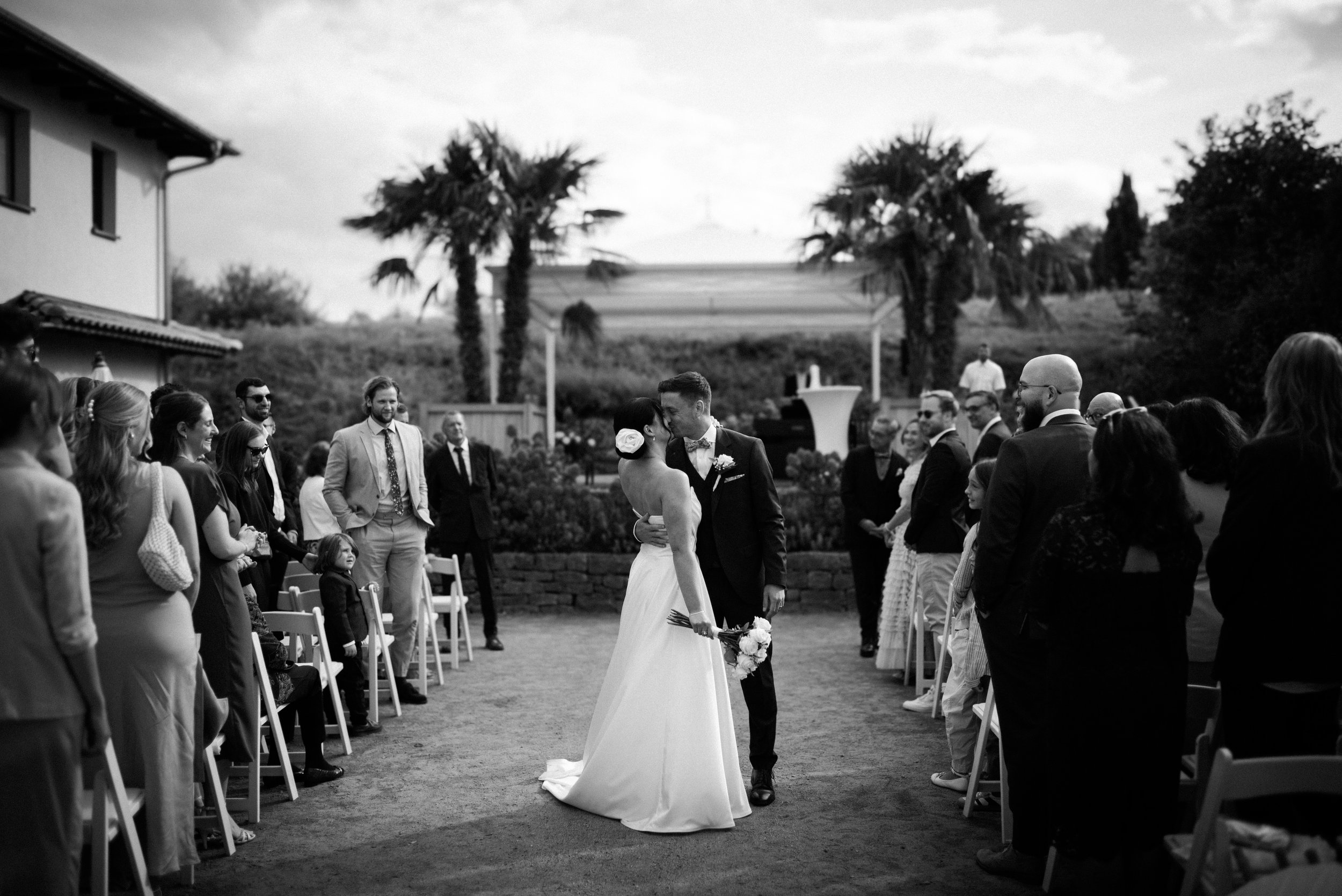 Braut und Bräutigam in der Mitte, in einem Garten bei einer Hochzeitsfeier, umgeben von Gästen, im Schwarz-Weiß-Stil.