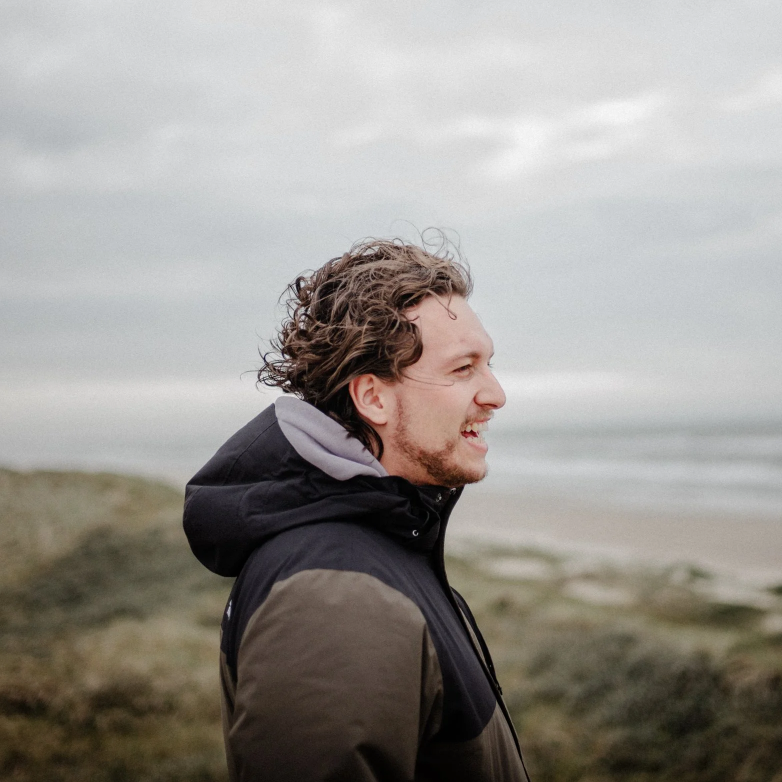 Ein lachender Mann mit lockigem Haar trägt einen schwarzen und grauen Mantel am Strand mit Felsen und dem Meer im Hintergrund bei bewölktem Himmel.