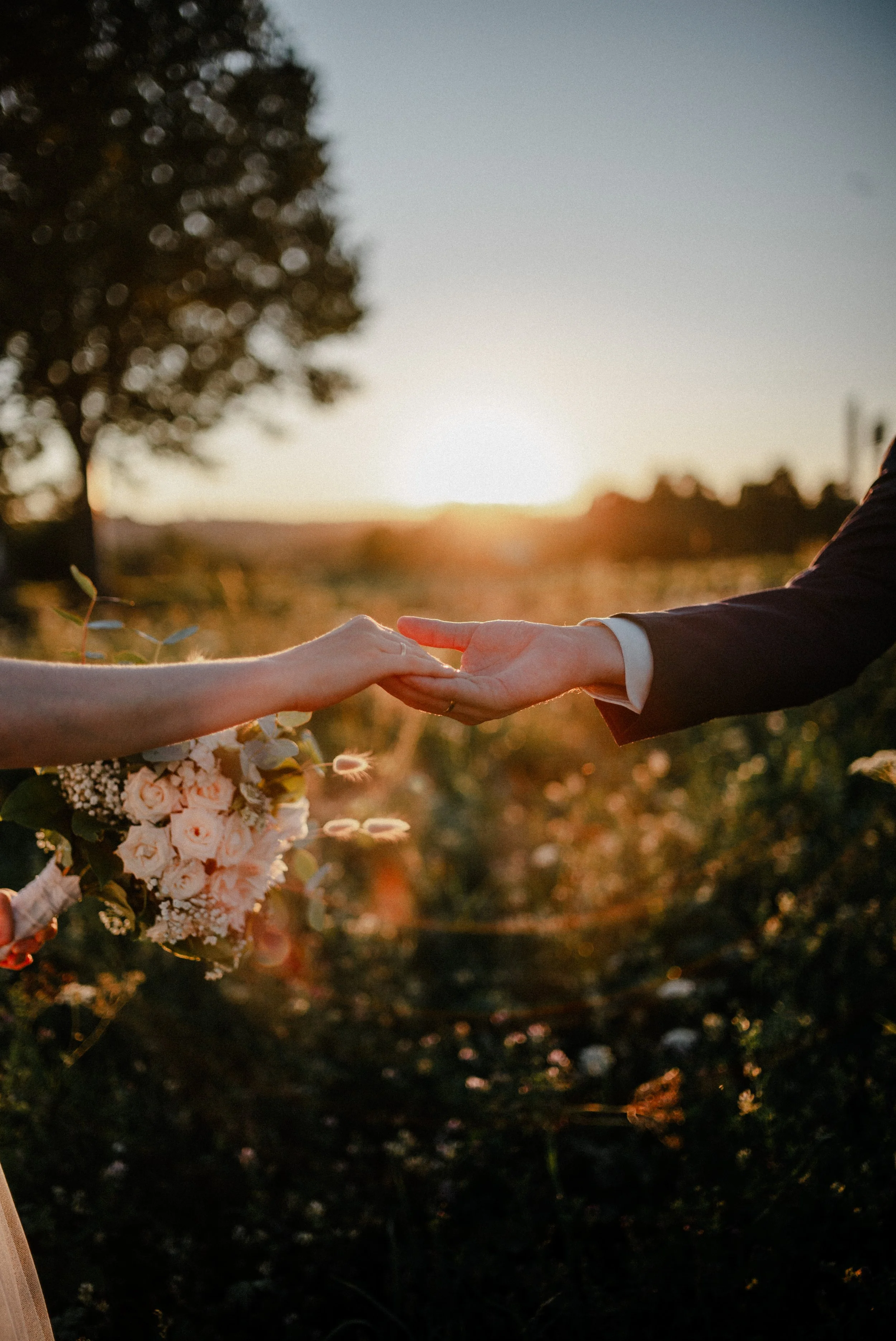Zwei Menschen berühren sich mit den Händen bei Sonnenuntergang im Freien, eine Person hält einen Blumenstrauß.