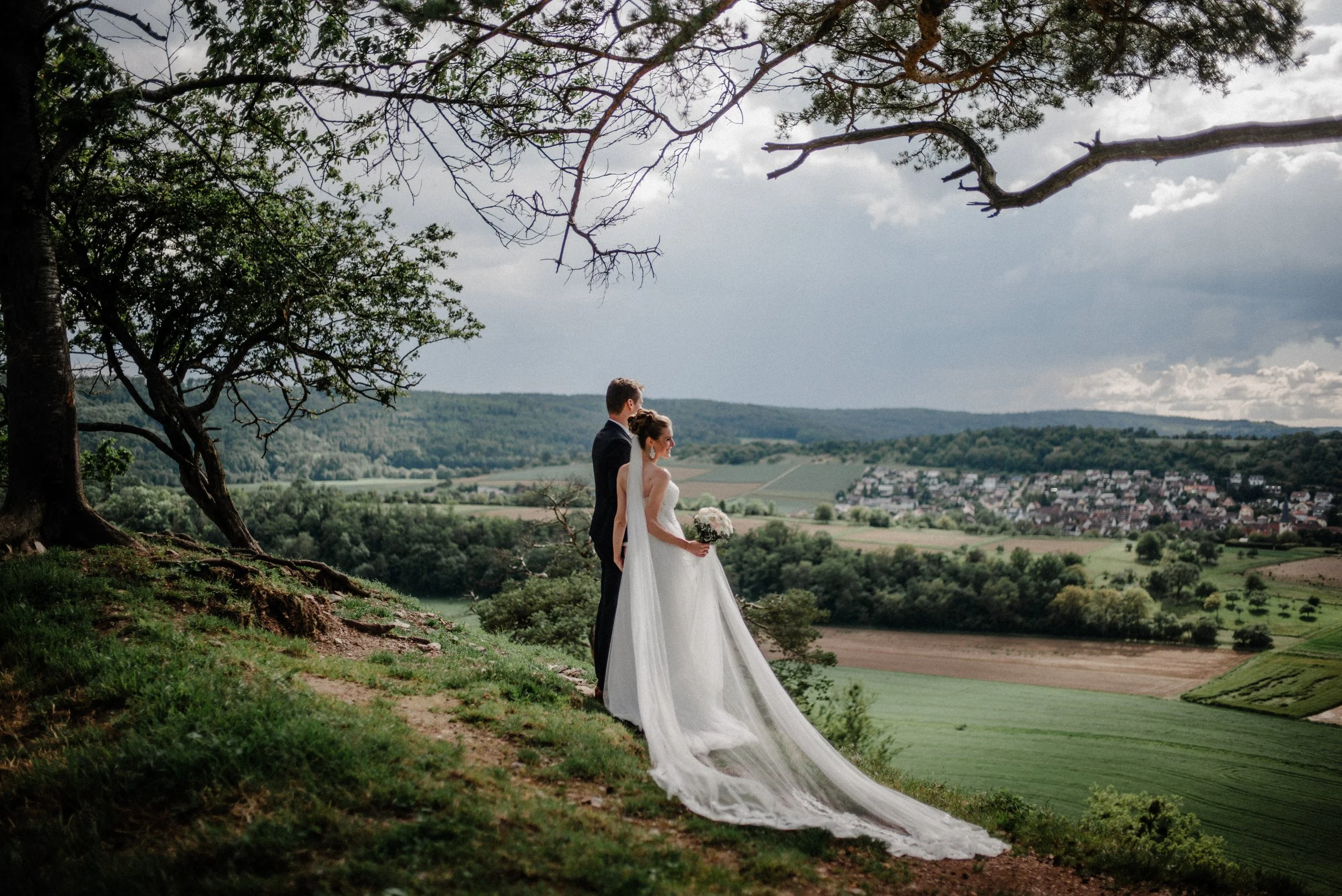 Ein Hochzeitsbild mit einem Brautpaar im weißen Kleid und Anzug, die auf einer Anhöhe mit Blick auf eine grüne Landschaft und eine kleine Stadt stehen, umgeben von Bäumen, bei bewölktem Himmel.