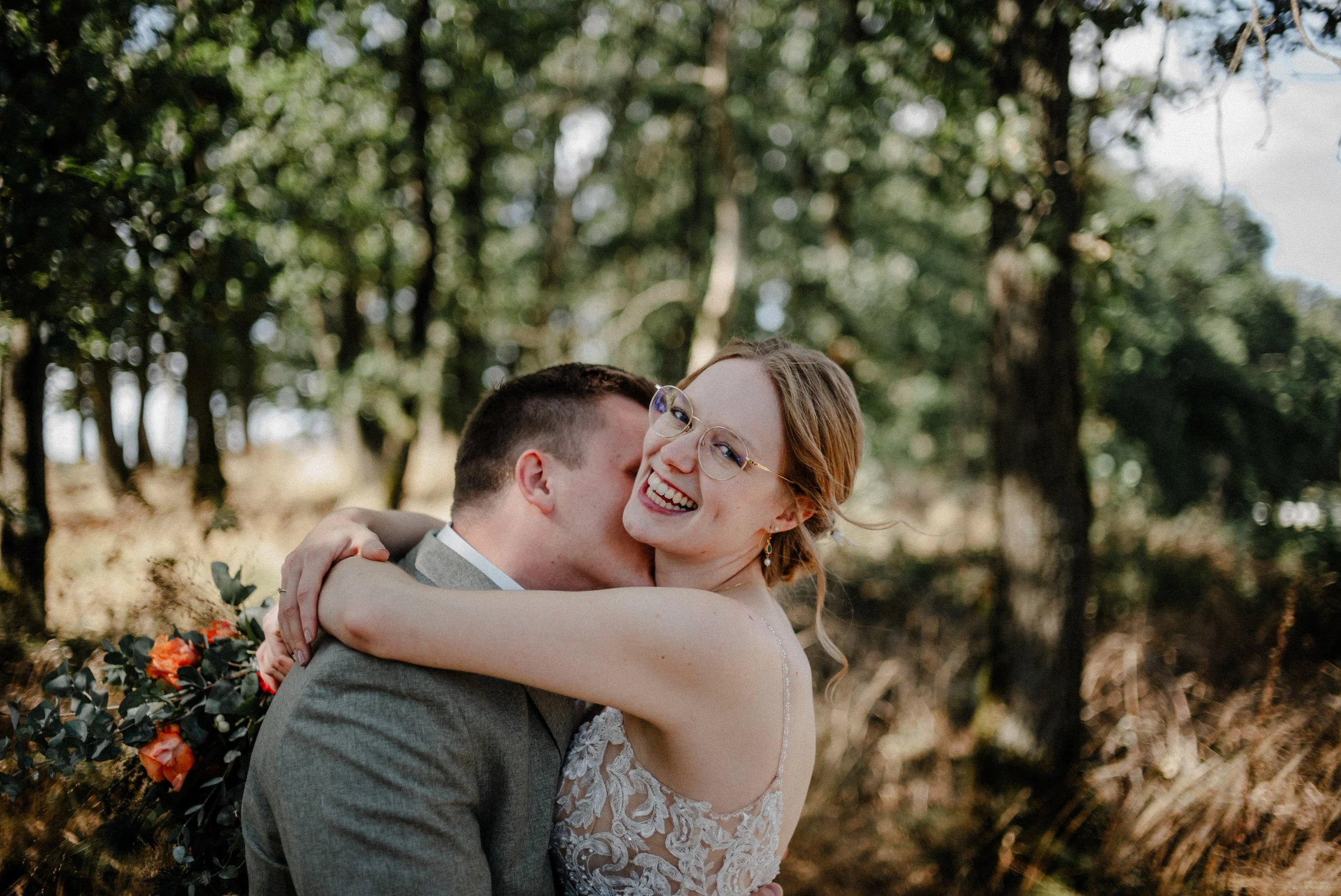 Ein glückliches Hochzeitspaar umarmt sich in einem Wald, das Brautpaar lacht, die Braut trägt eine Spitzenhochzeitskleid mit Spaghettiträgern, der Bräutigam trägt einen grauen Anzug.