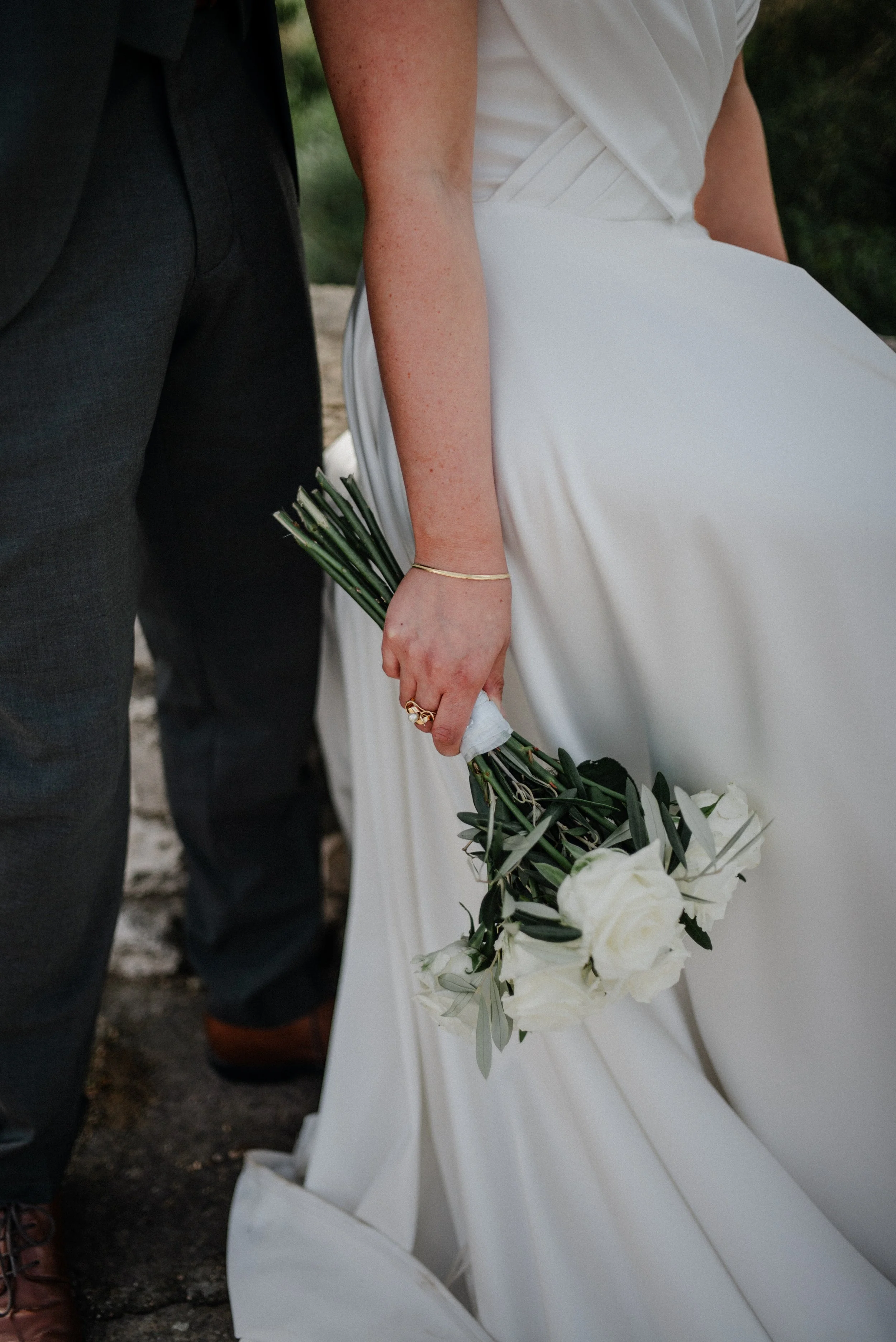 Naheaufnahme eines Brautpaares, bei der die Braut ein Bouquet aus weißen Rosen und grünen Blättern hält, während sie in einem Hochzeitsoutfit sitzt.