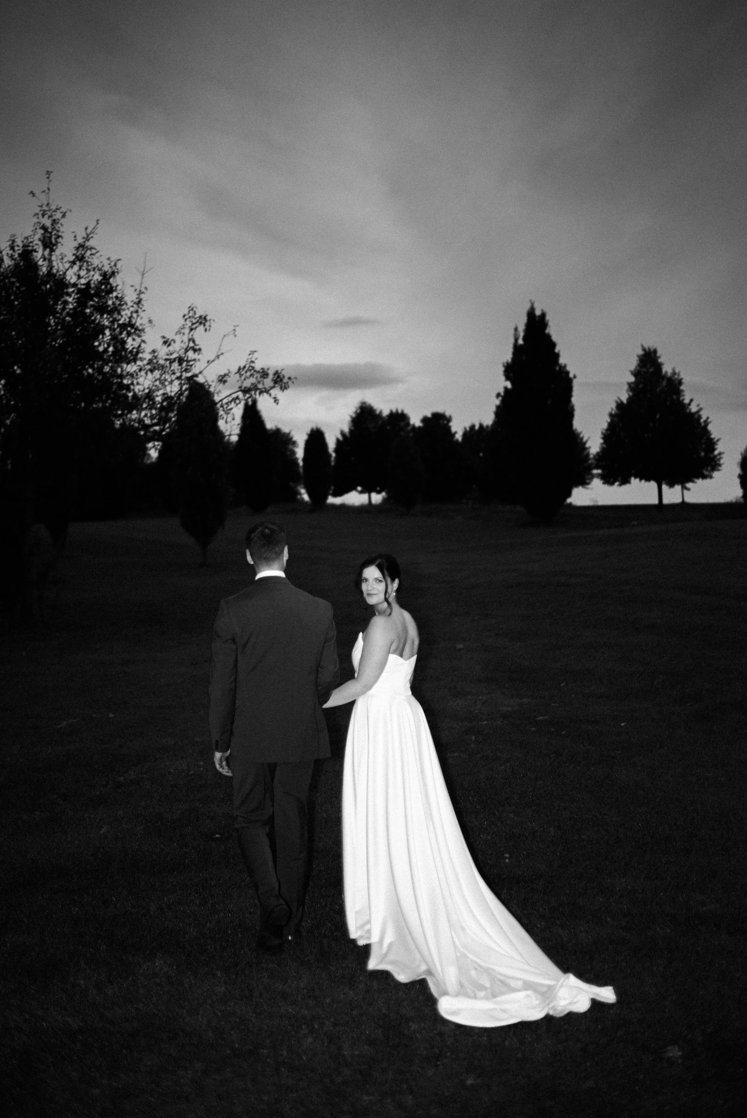 Ein Brautpaar läuft Hand in Hand auf einem Rasen im Freien bei Sonnenuntergang, mit Bäumen im Hintergrund und einer dunklen Himmelsszene.