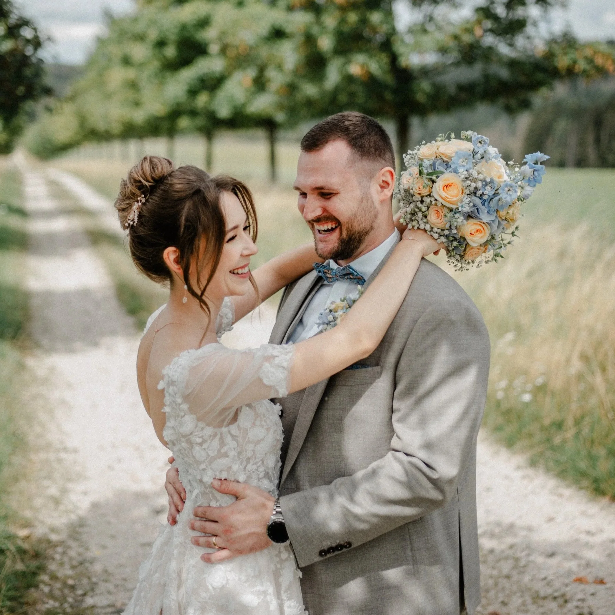Ein verliebtes Brautpaar in einem Feld, lachend und umarmend, mit einem Blumenstrauß bei einem Hochzeitsfoto.