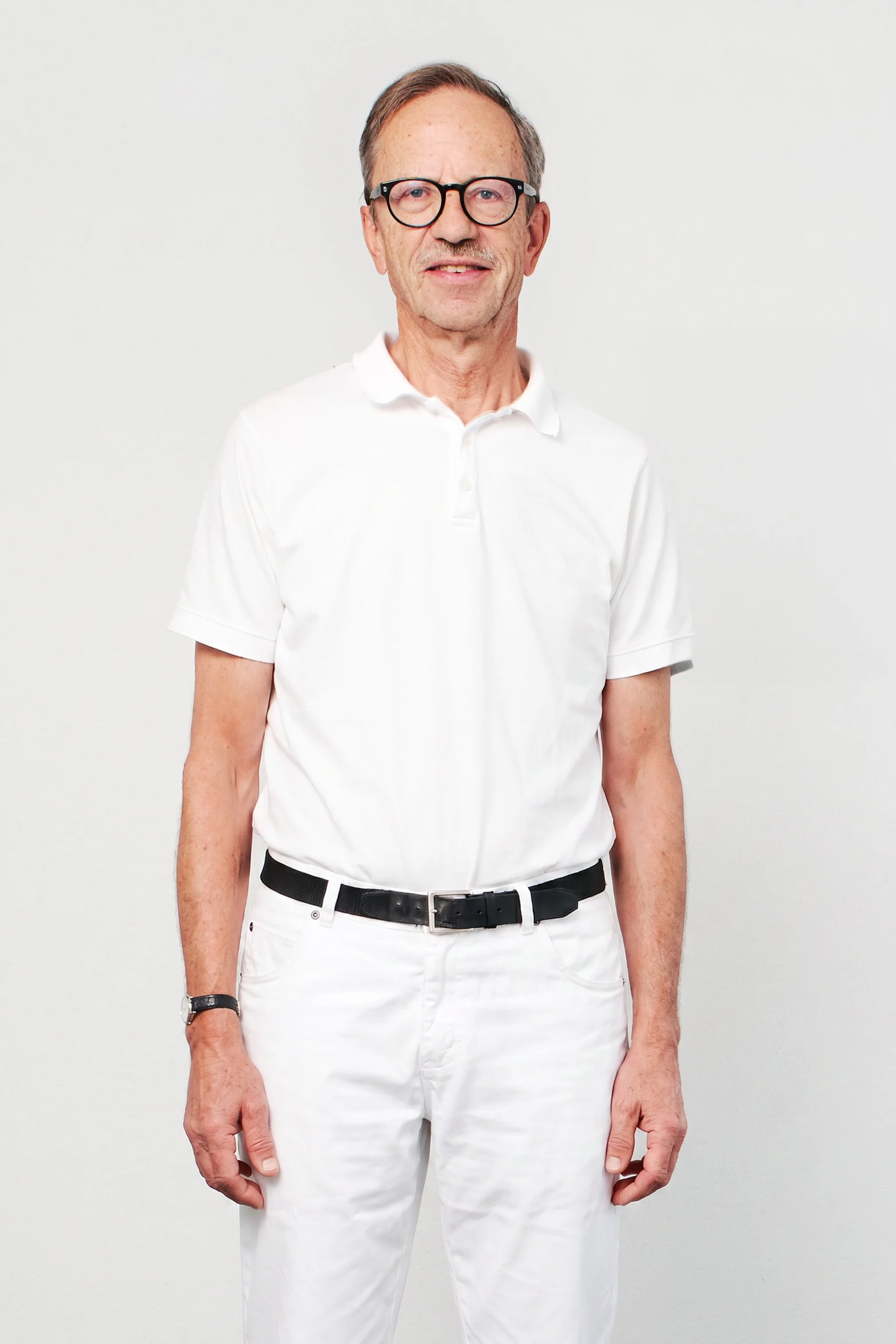 Älterer Mann mit Brille in weißem Hemd und weißen Hosen vor weißem Hintergrund.
