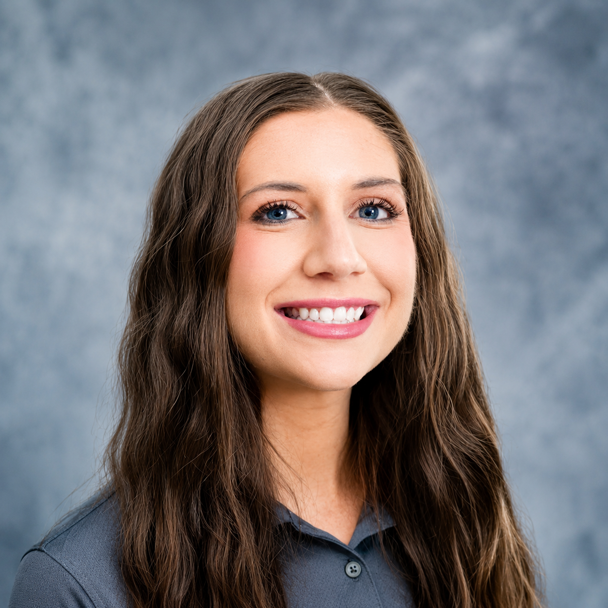 A young woman with long brown hair, smiling with white teeth, wearing a dark collared shirt, against a grey textured background.