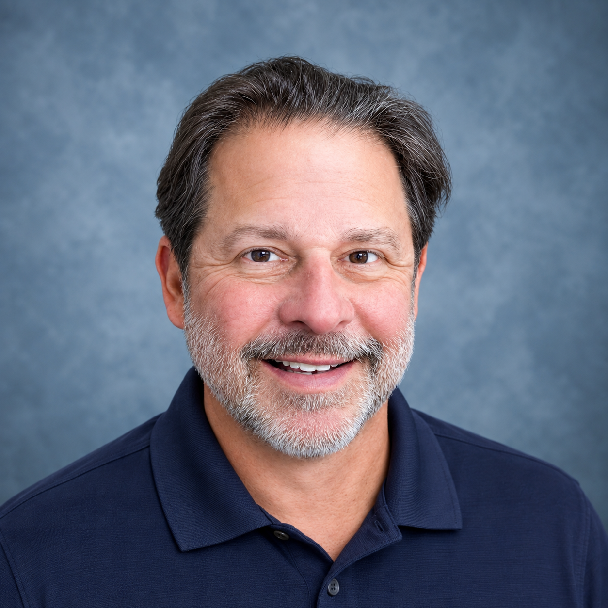 A middle-aged man with a beard and mustache, smiling, wearing a checkered shirt and a dark jacket, against a gray background.