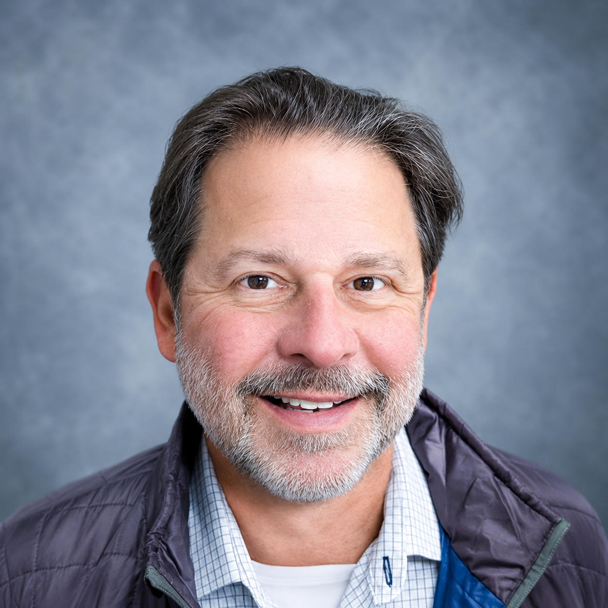 A middle-aged man with a beard and mustache, smiling, wearing a checkered shirt and a dark jacket, against a gray background.