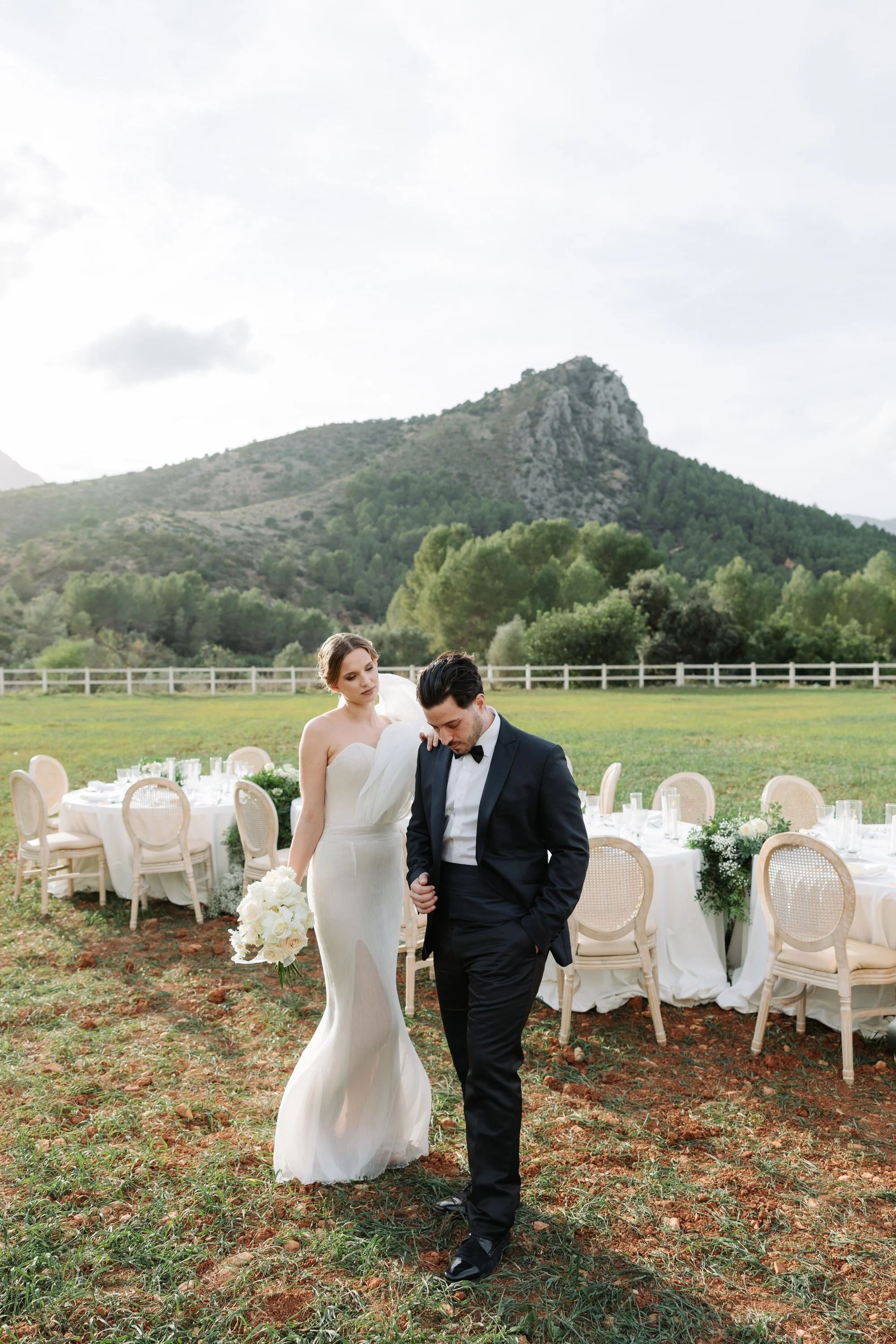 Pareja de novios en ceremonia al aire libre, la novia con vestido blanco y el novio con saco negro, en un entorno natural con mesas decoradas y montañas en el fondo.