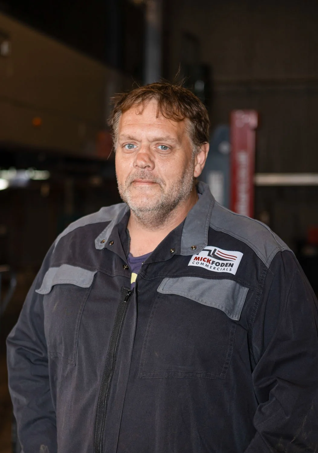 Man standing outdoors near a vehicle with yellow and red stripes, wearing a dark jumpsuit and maroon beanie.
