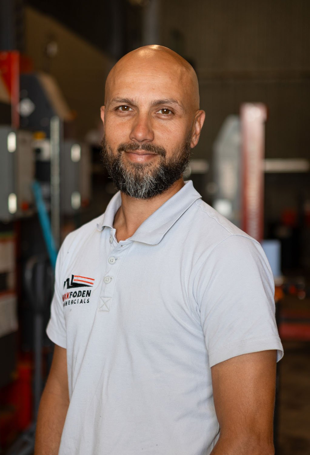 A man with a beard and bald head wearing a gray zip-up jacket, standing in front of a textured, cracked white wall.