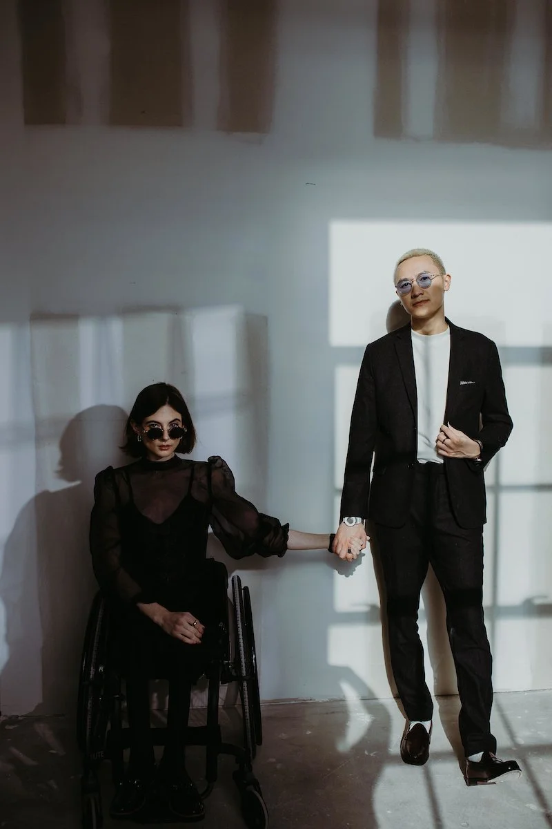 Man in suit holding hand of woman in wheelchair, both wearing sunglasses, against a shadowed wall.