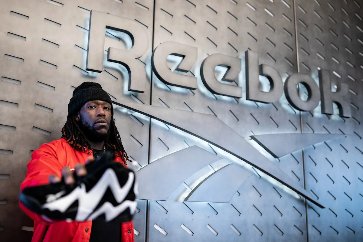 Person in red jacket holding Reebok sneaker in front of Reebok logo.