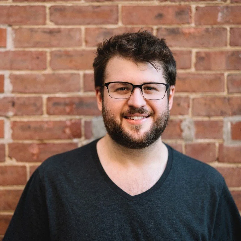Person with glasses and beard smiling in front of a brick wall.