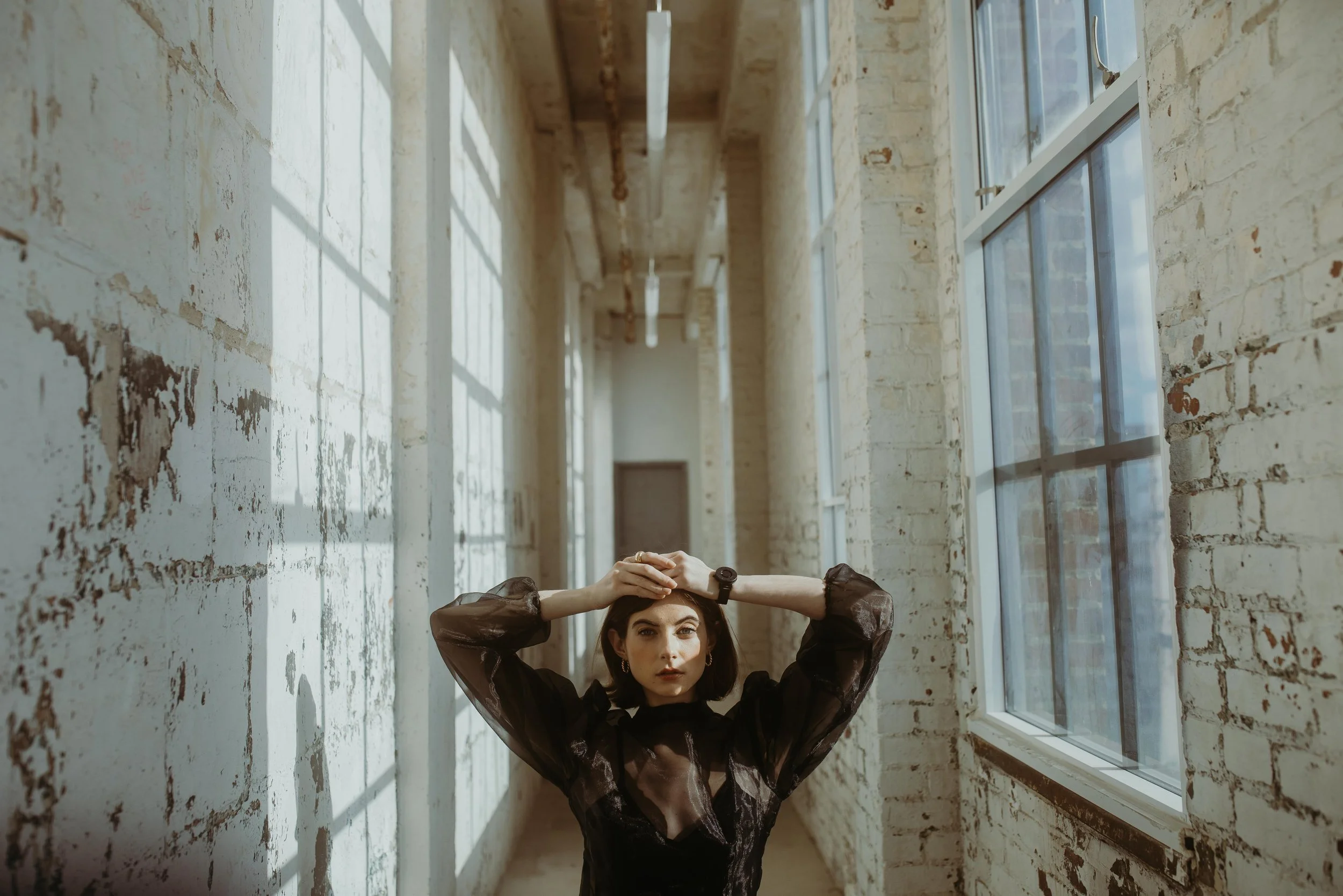 Person standing in a narrow hallway with brick walls and windows, wearing a black top, hands resting on their head.